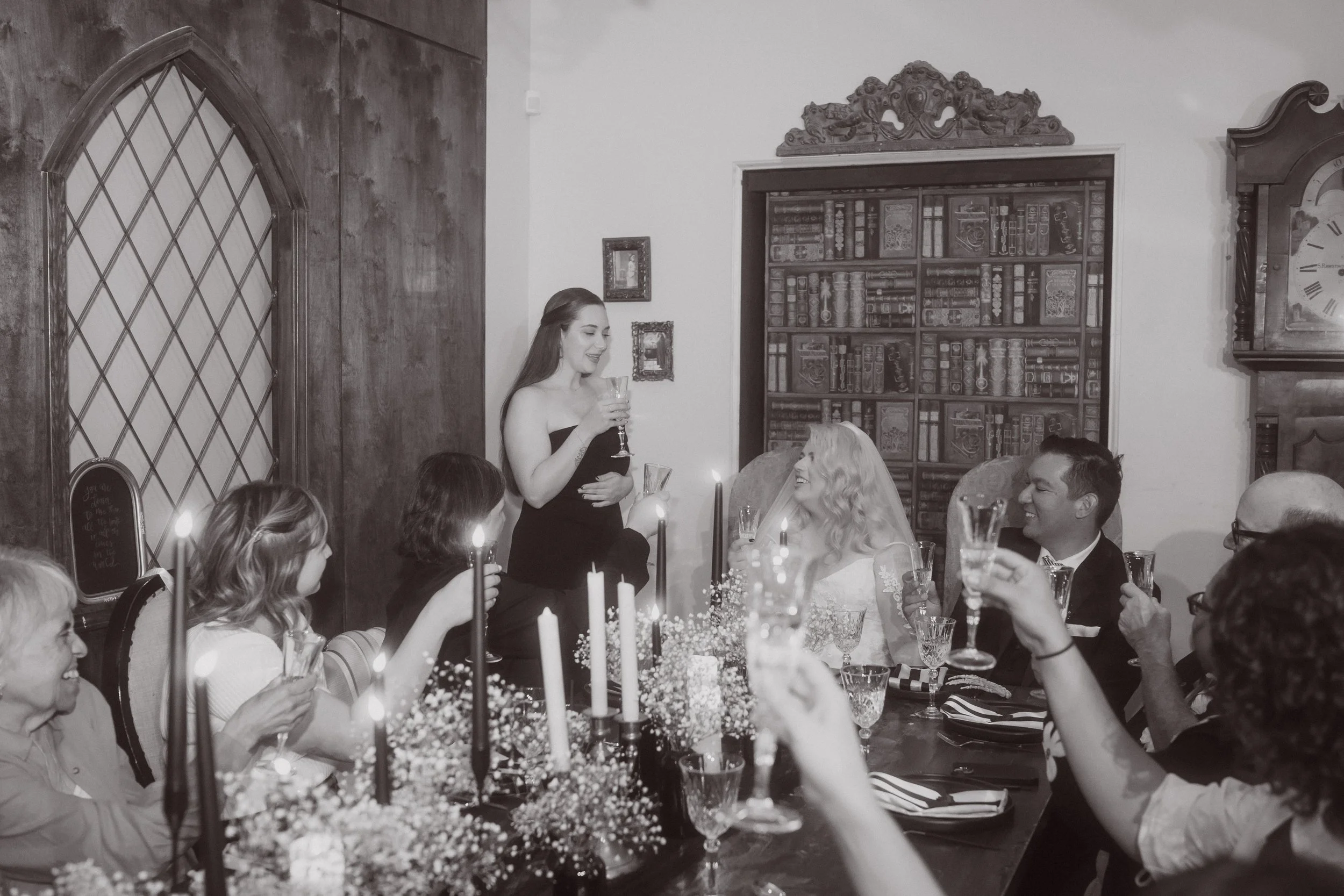 Wedding guest giving a toast at an intimate candlelit reception with long table, black taper candles, and floral centerpieces