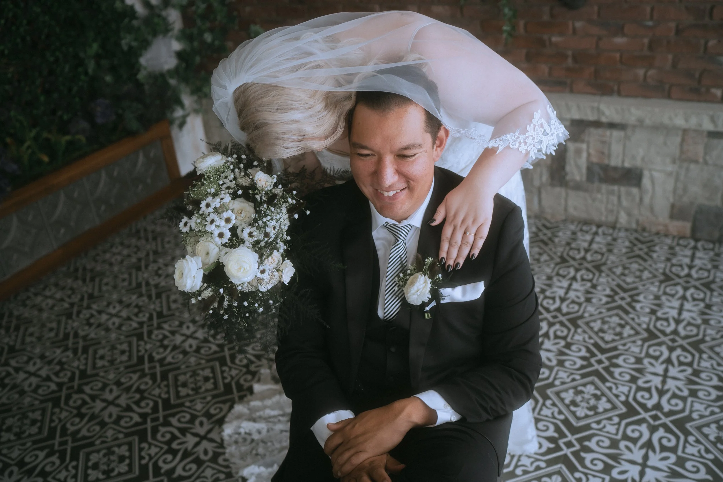 Bride leaning over groom with bouquet in intimate wedding portrait on patterned tile floor with greenery backdrop