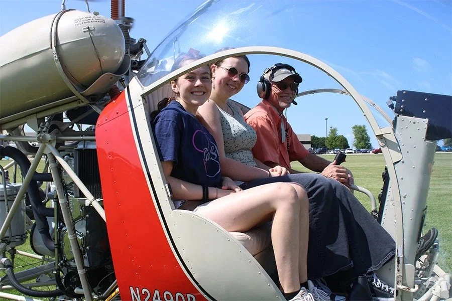 A kid, their parent, and a pilot smile from the open cockpit of a helicopter