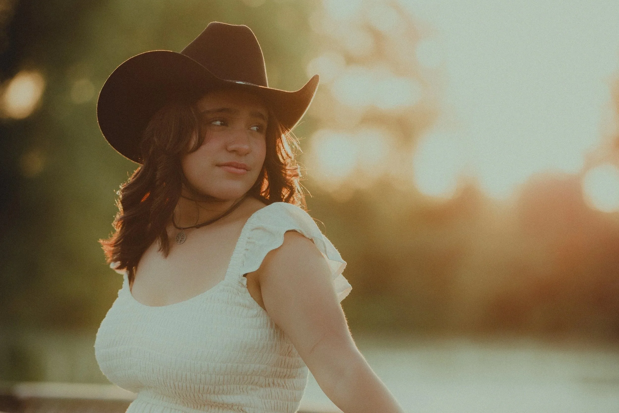 A young woman wearing a wide-brimmed cowboy hat and a white dress, looking to the side, with a blurred outdoor background illuminated by warm sunset light.