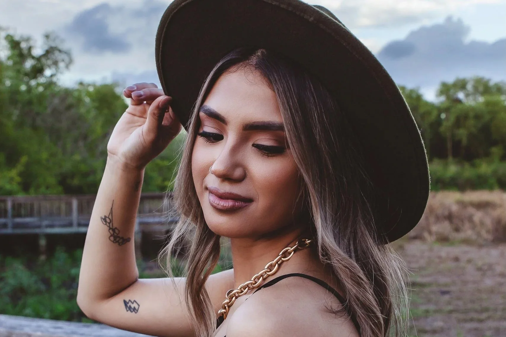A woman with tattoos on her arm and wearing a black spaghetti strap top, gold chain necklace, and a wide-brimmed hat, standing outdoors with greenery and a cloudy sky in the background, looking down with a slight smile.
