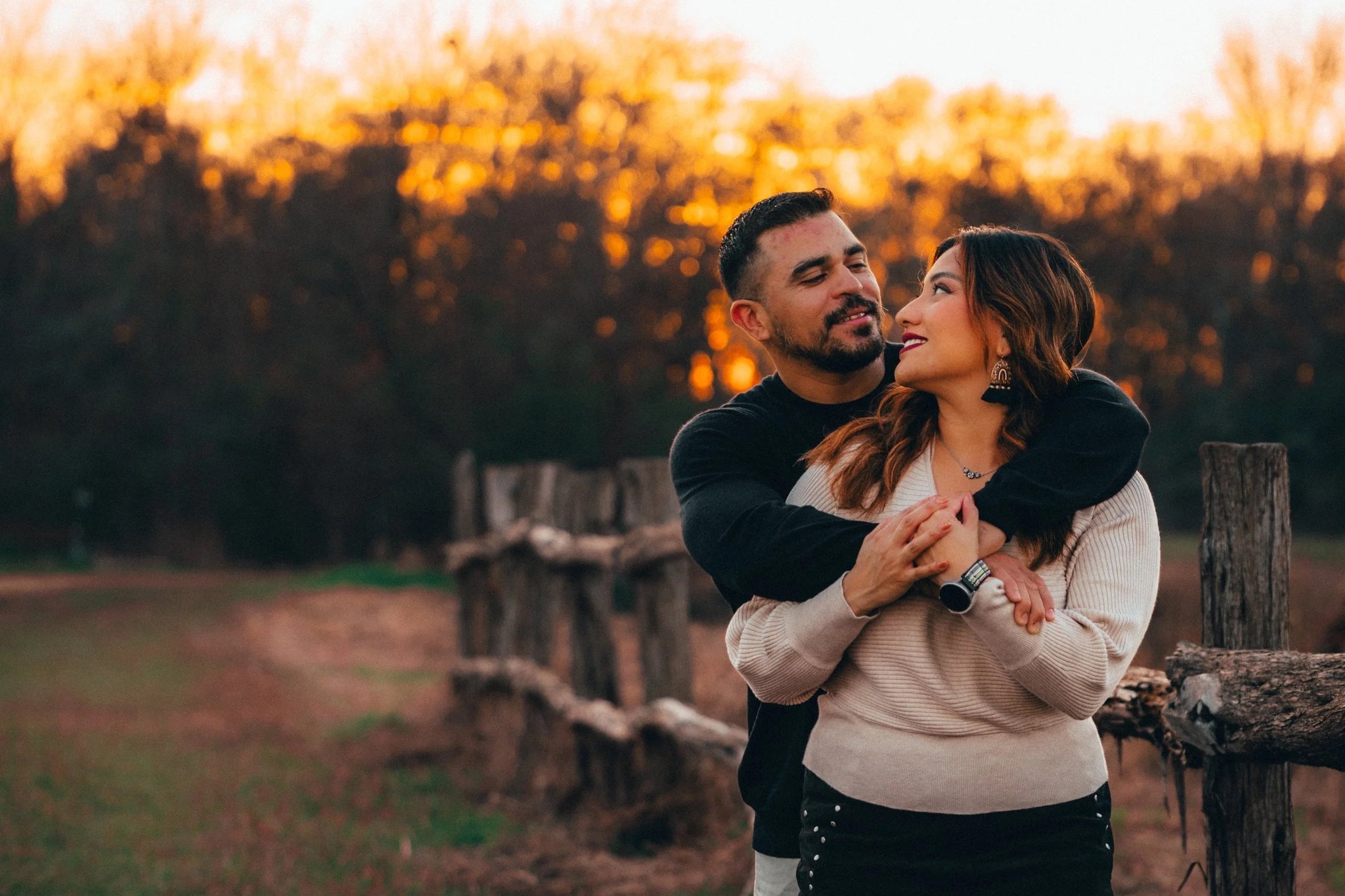 A couple embracing outdoors during sunset, with man hugging woman from behind and both looking at each other affectionately.