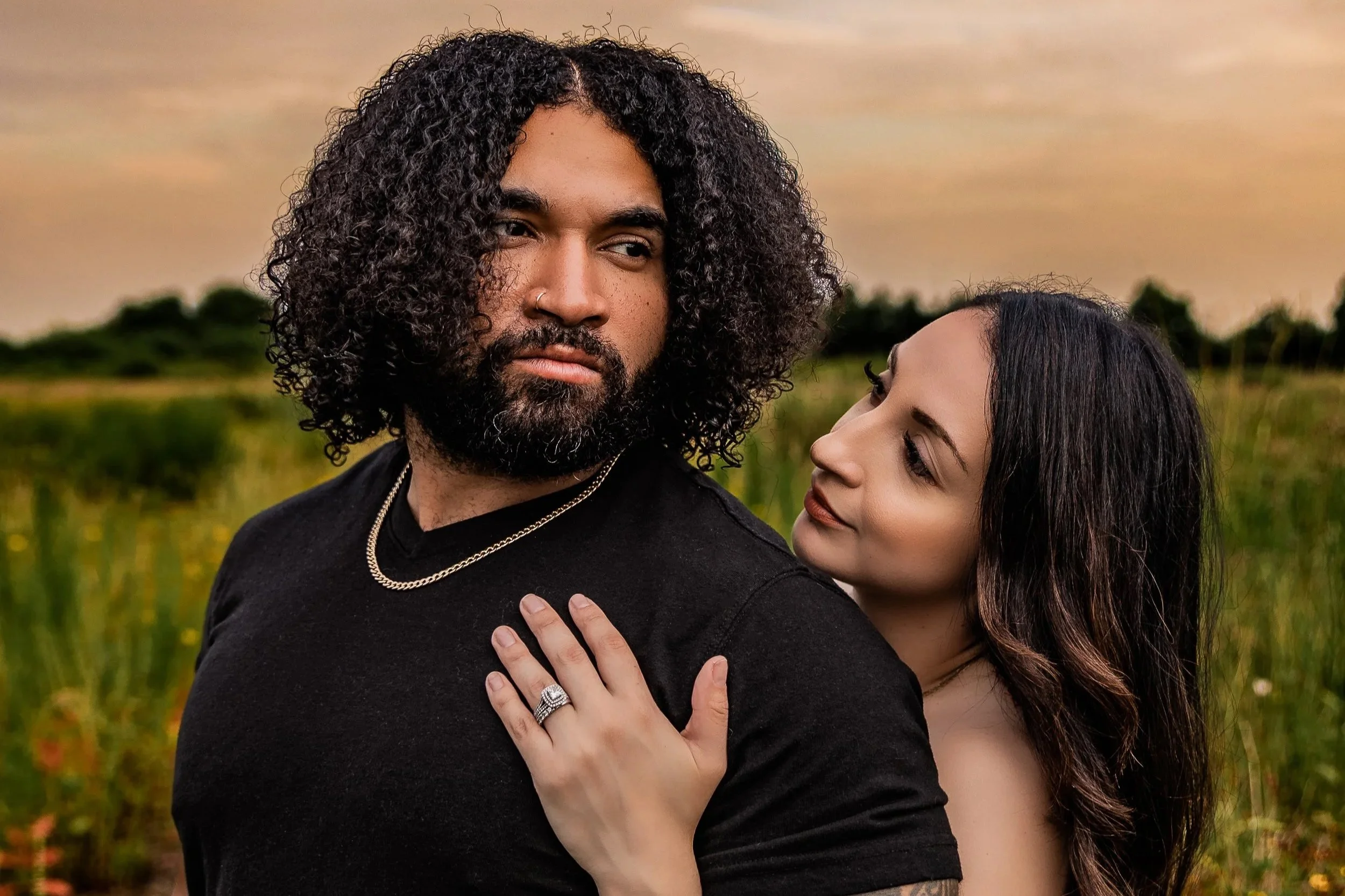 A man with curly hair and a beard, wearing a black shirt and jewelry, is looking to the side as a woman with long dark hair leans her head on his shoulder, gazing at him in a field during sunset.