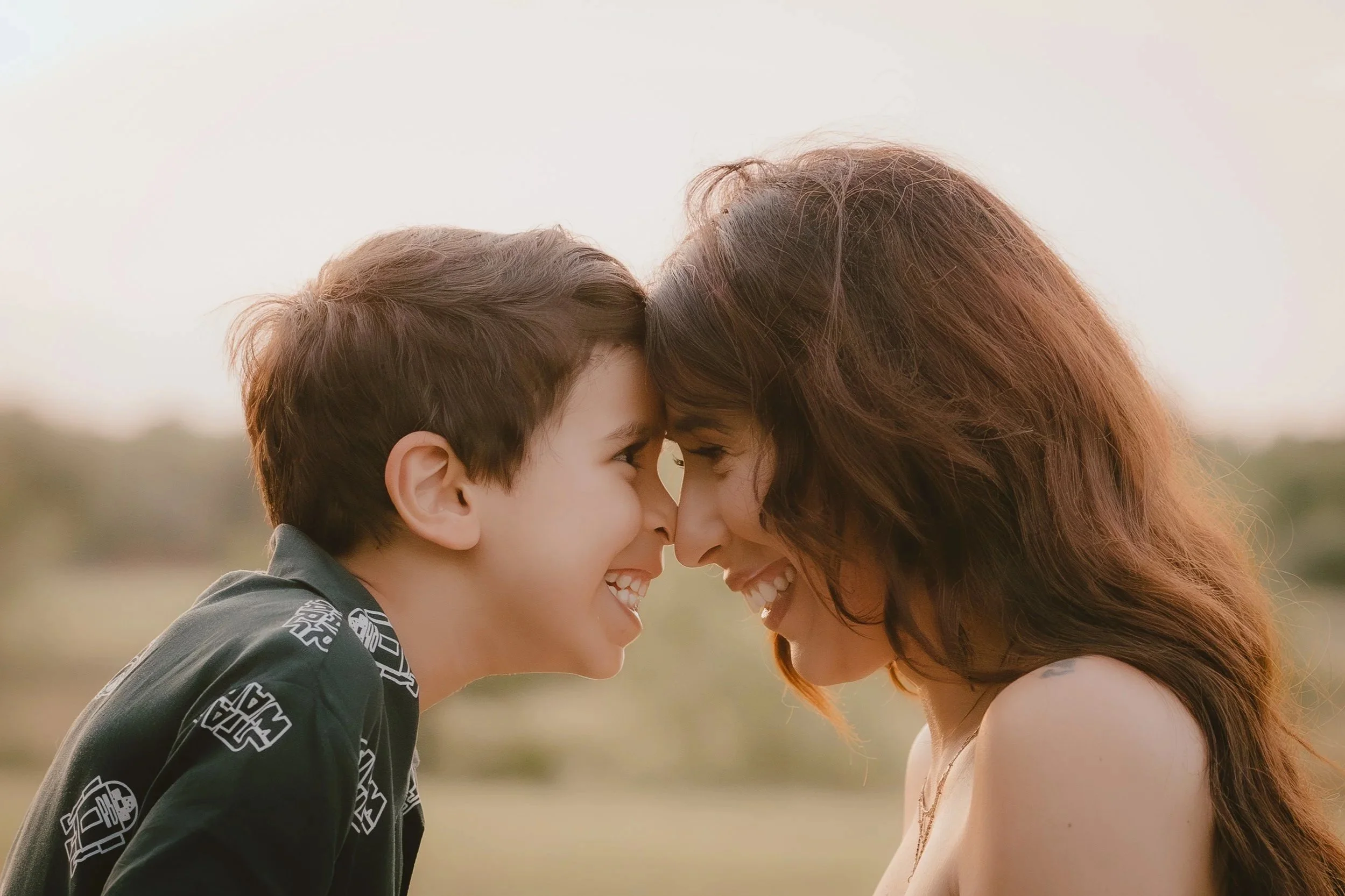 A boy and woman are smiling with their foreheads touching outdoors on a cloudy day.