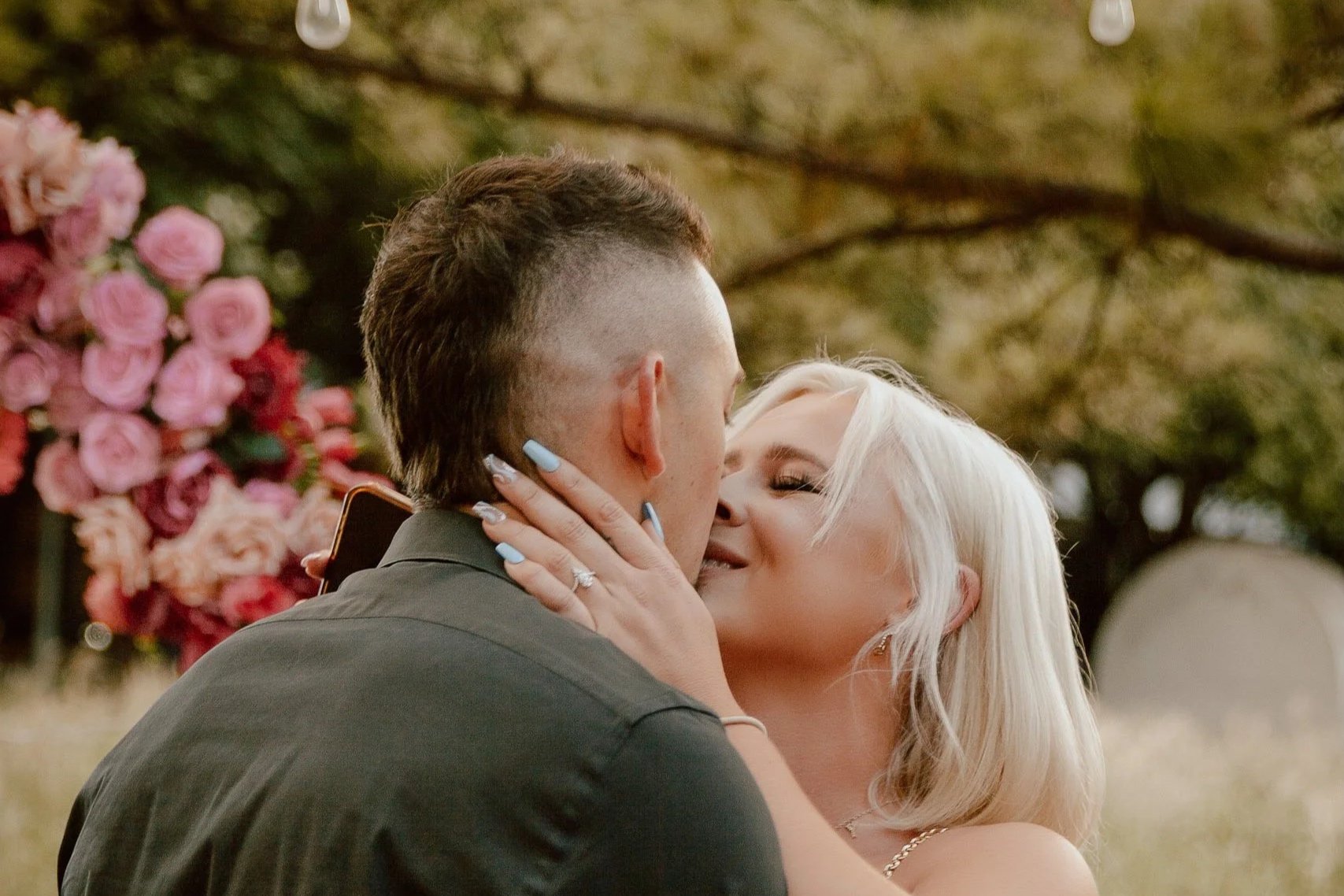 A couple sharing a kiss outdoors with pink flowers and green foliage in the background.
