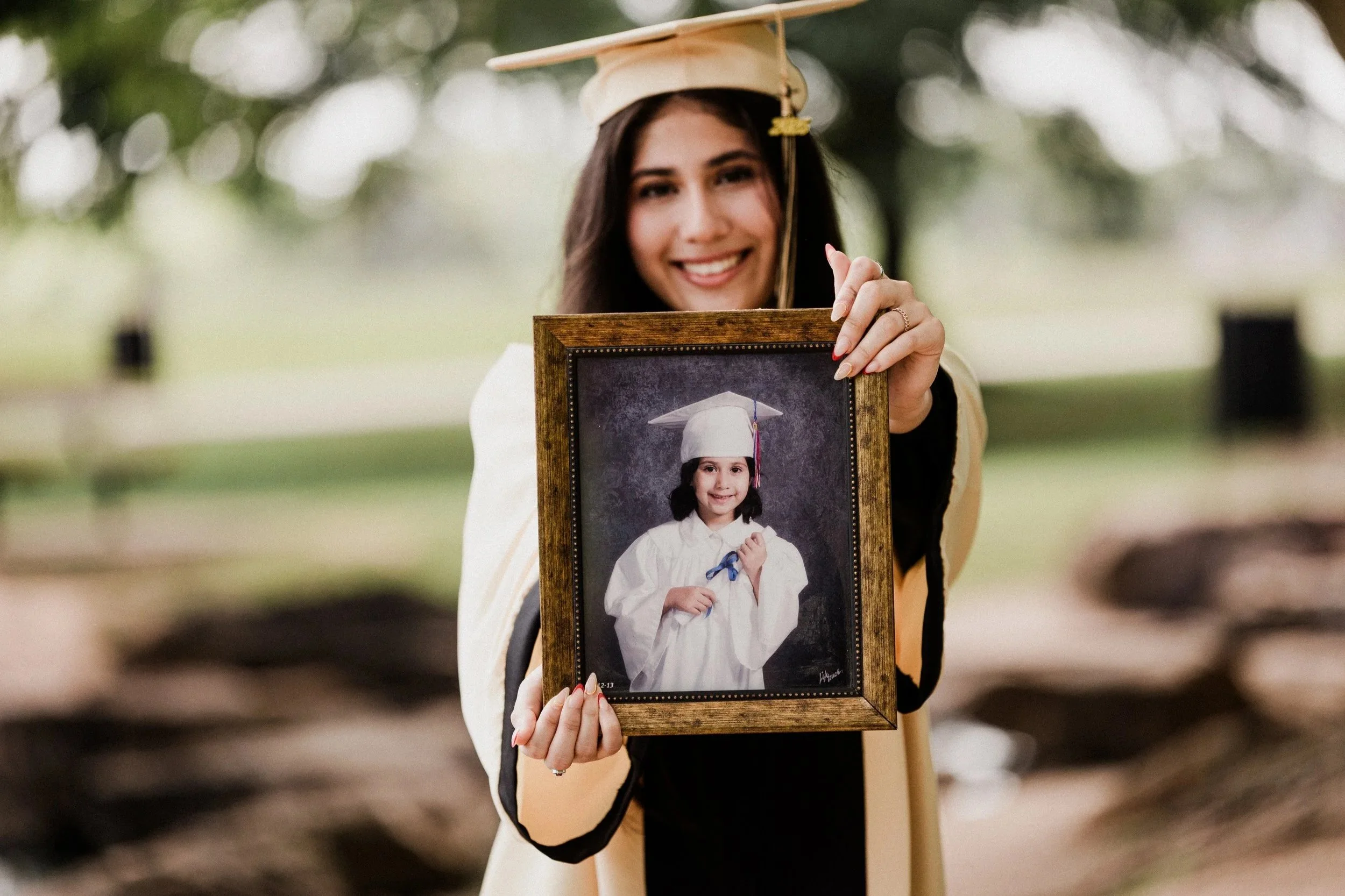 A woman in a graduation cap and gown holding a framed photo of herself as a young girl in graduation attire, outdoors with trees in the background.