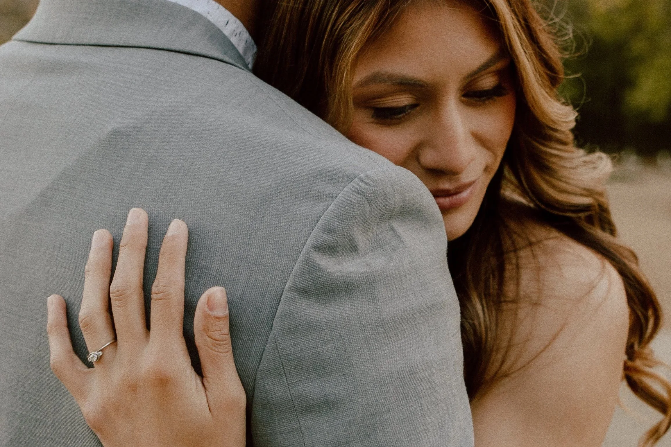 A woman embracing a man, her hand resting on his shoulder, with a wedding ring visible. She has long, wavy hair and a tender expression.