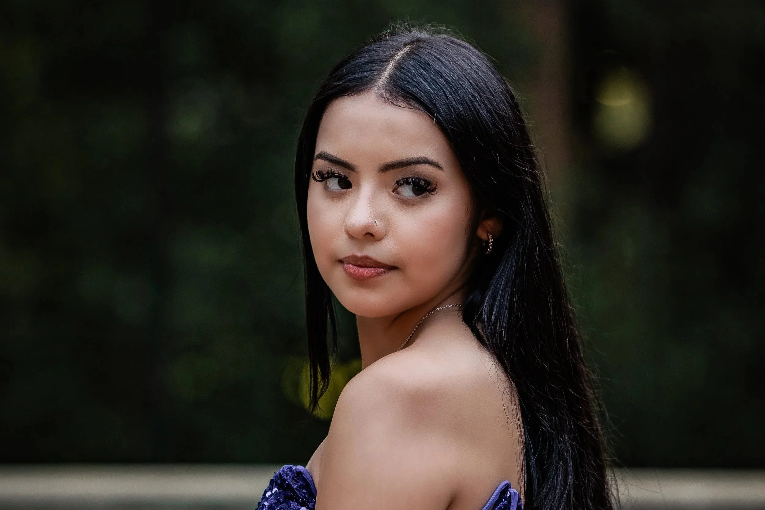 A young woman with long black hair, wearing earrings and a necklace, looking over her shoulder outdoors with a blurred natural background.