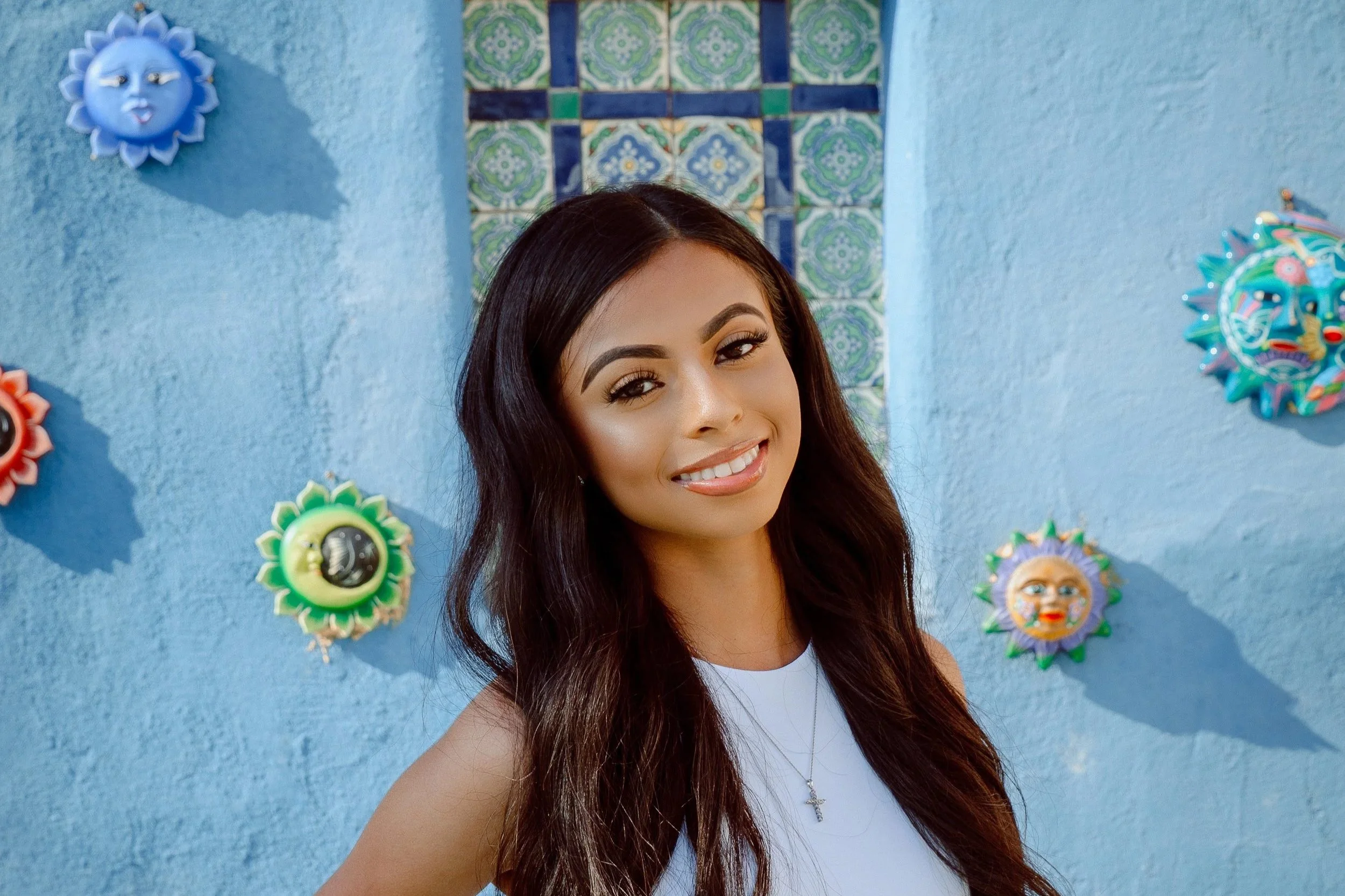 A young woman with long dark hair smiling in front of a blue wall decorated with colorful sun and moon masks and tiles.