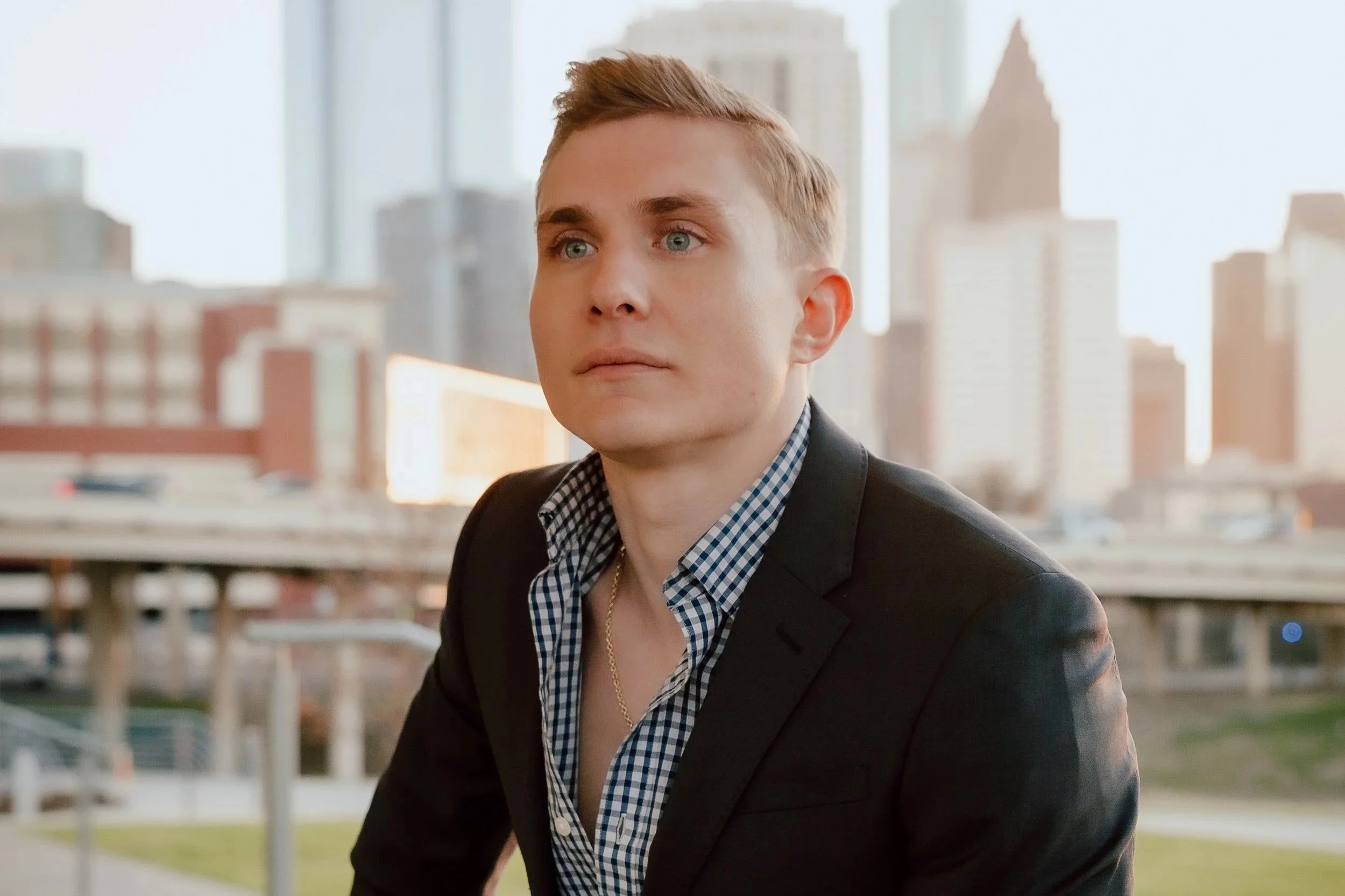 A young man with blonde hair, blue eyes, wearing a black blazer over a blue and white checkered shirt, sits outdoors with a city skyline in the background during daylight.