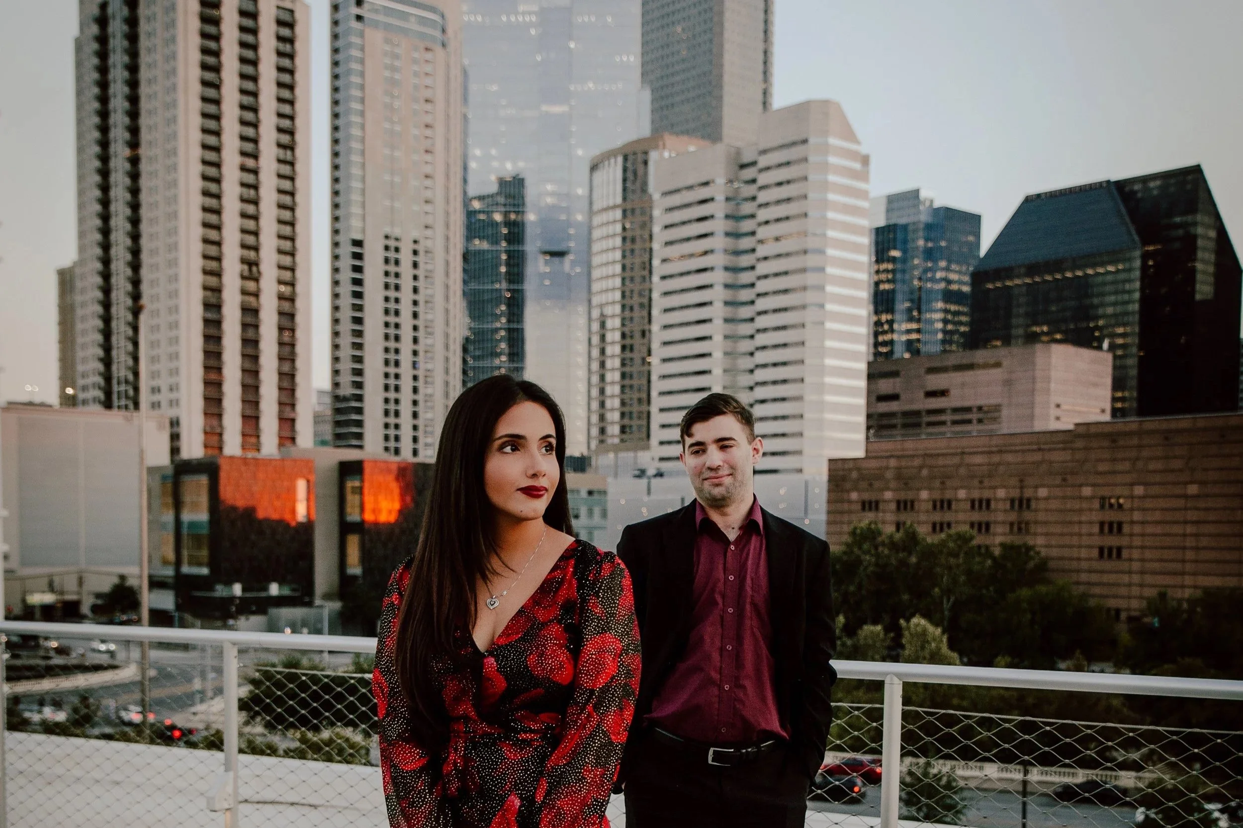 A woman in a red and black floral dress and a man in a dark suit with a red shirt standing on a rooftop with a city skyline in the background during dusk.