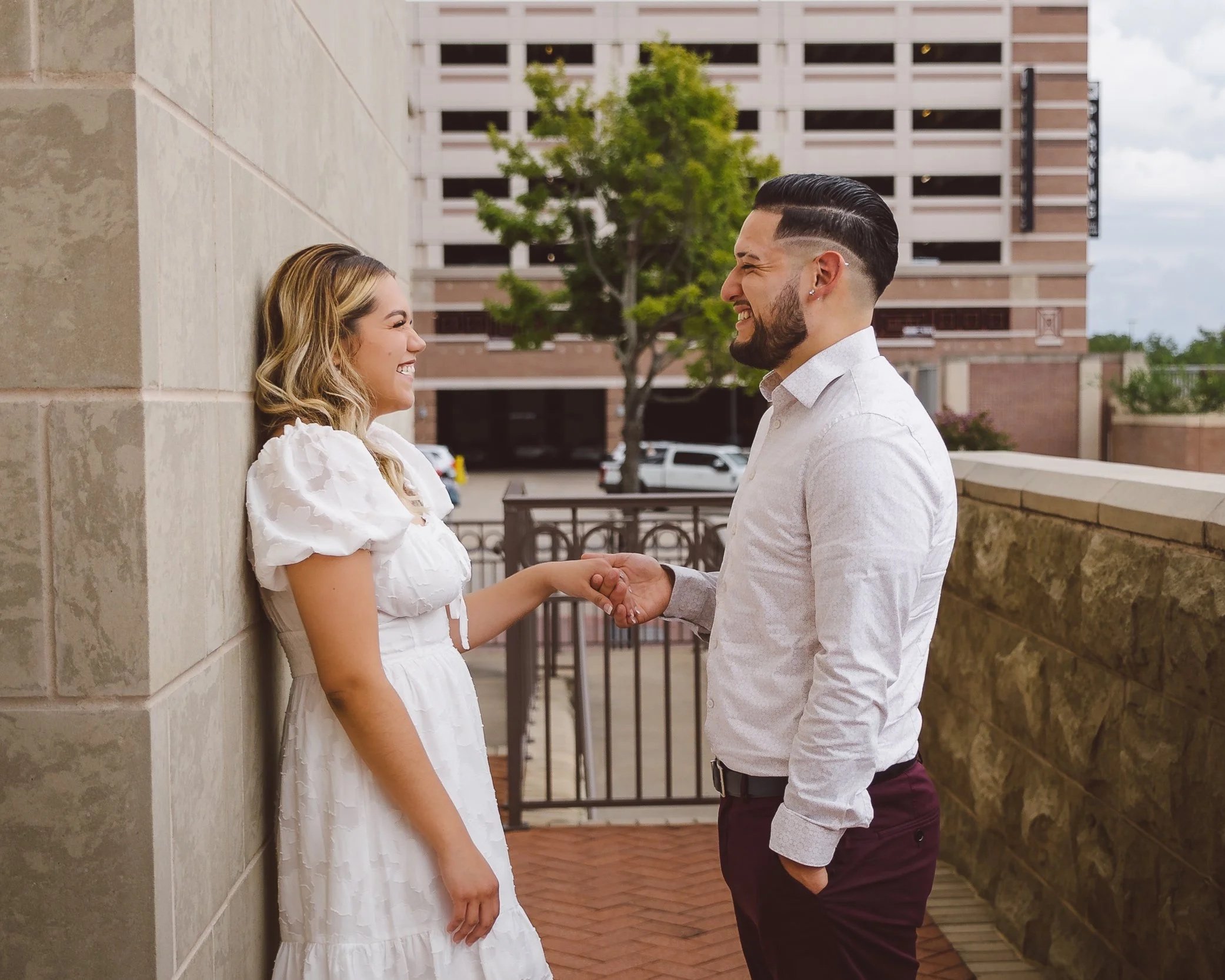 A woman in a white dress and a man in a white shirt and dark pants hold hands and smile at each other while standing outside near a stone wall with a cityscape in the background.