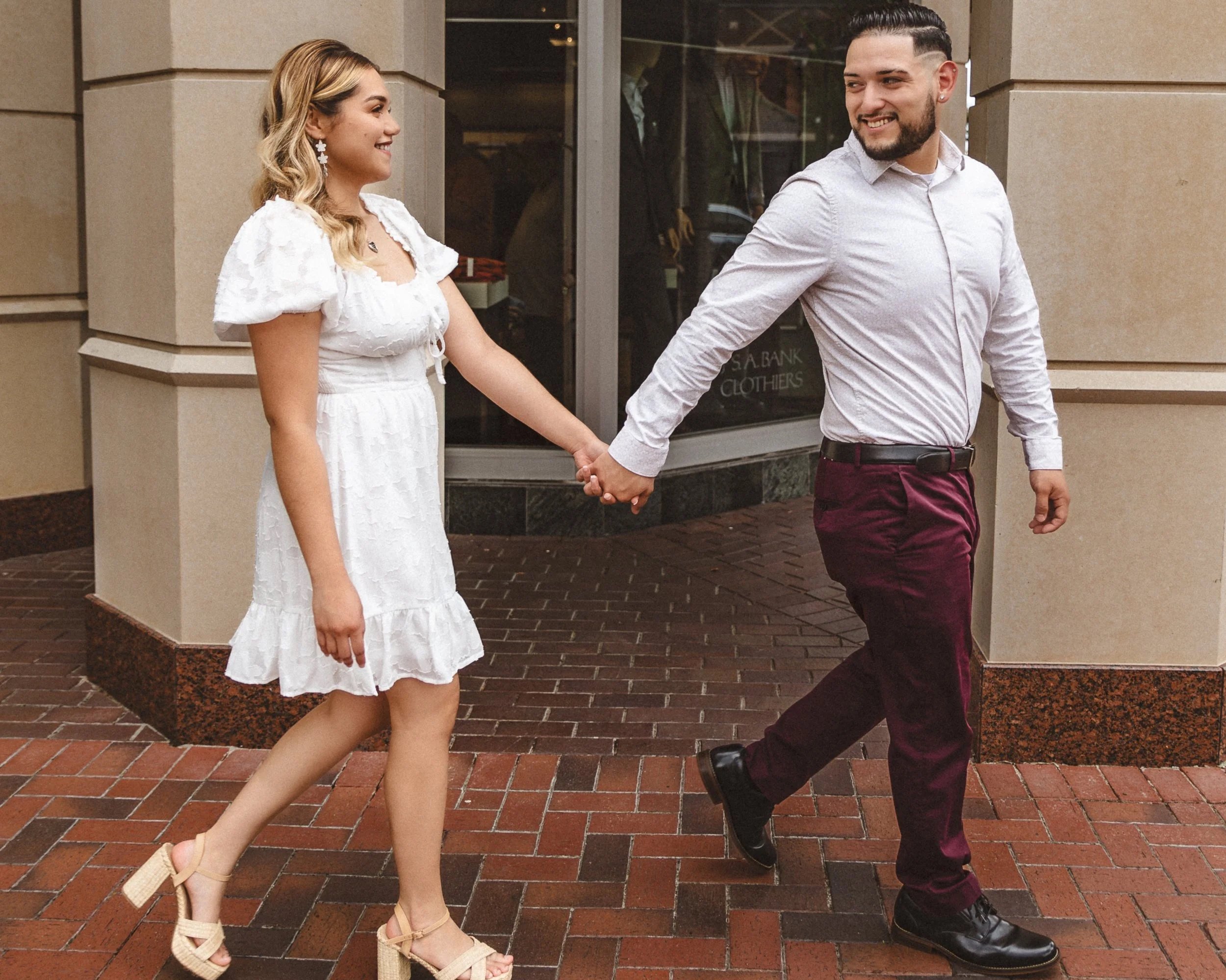 A couple holding hands and walking on a brick sidewalk outside a building, smiling at each other.