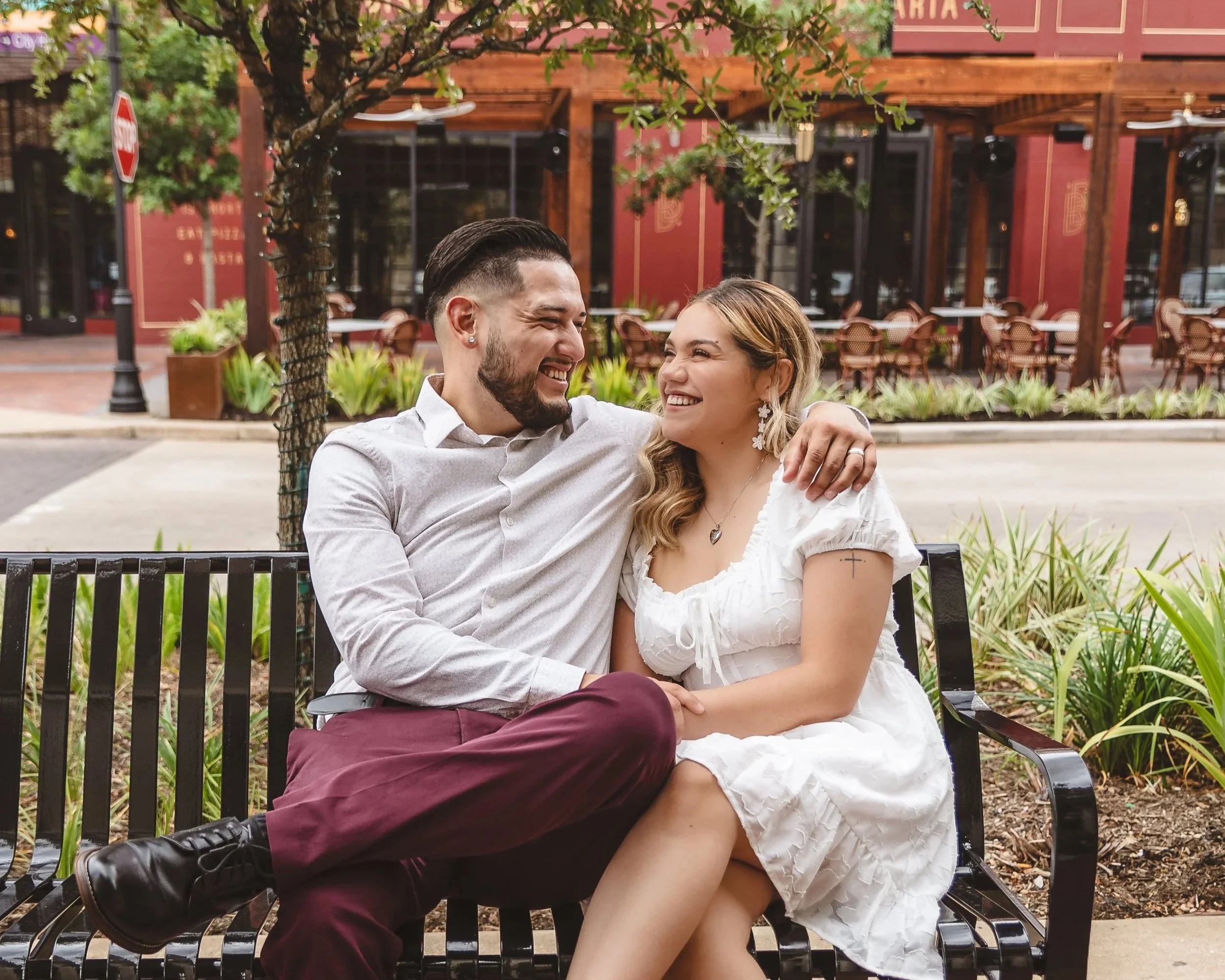 A happy couple sitting on a park bench, smiling and looking at each other. The man is wearing a light shirt and maroon pants, while the woman is in a white dress with jewelry. They are outdoors with greenery and a red restaurant building in the backg