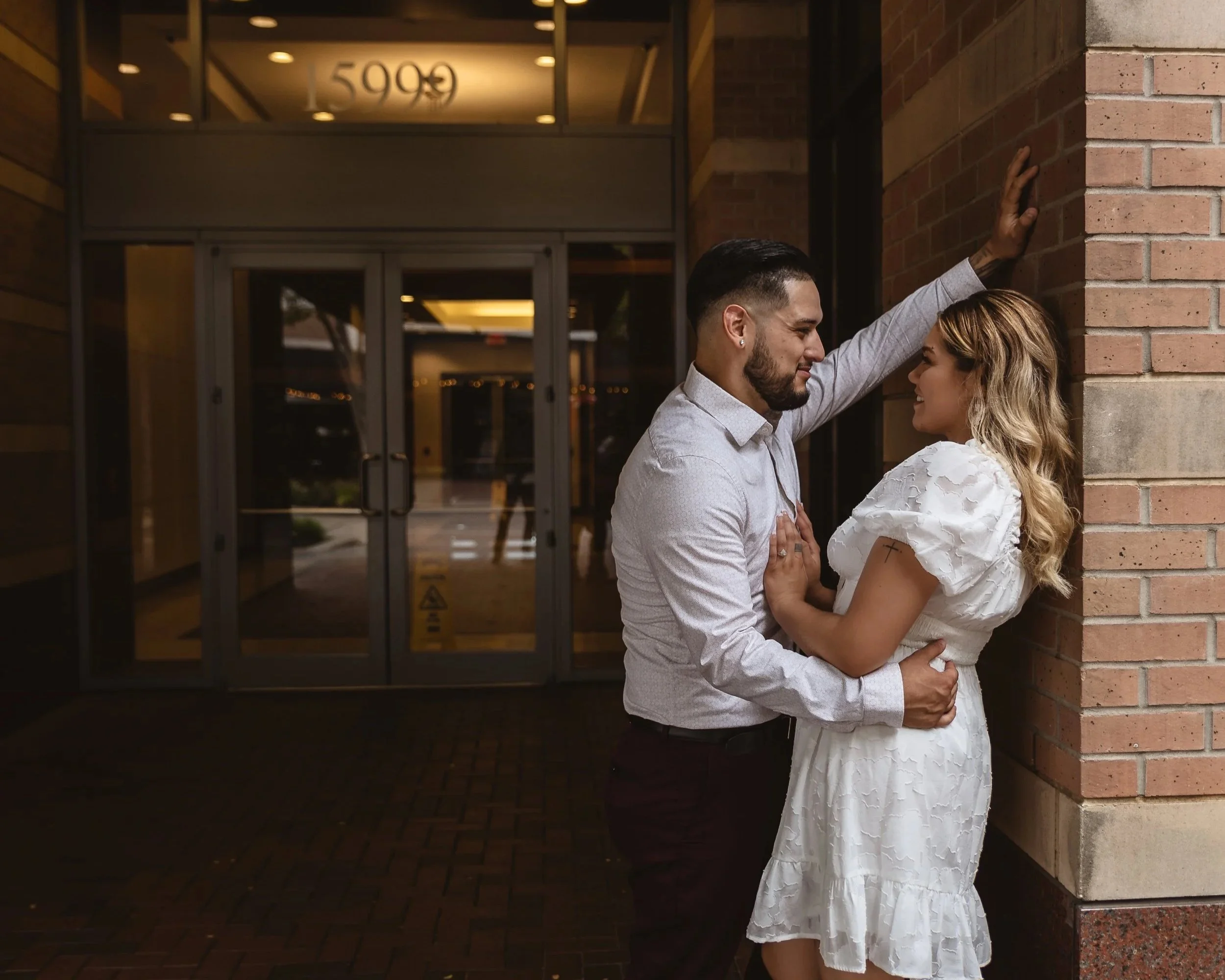 A couple standing close together outside a building at night; the man has his hand against the wall and the woman touches his chest, both smiling at each other.
