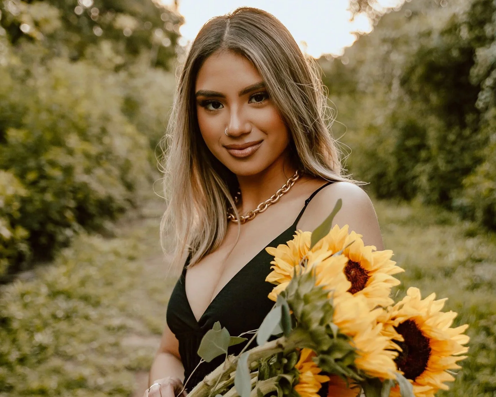 A woman with wavy hair wearing a black dress and a gold chain necklace, holding a bouquet of sunflowers, smiling outdoors during sunset with greenery in the background.