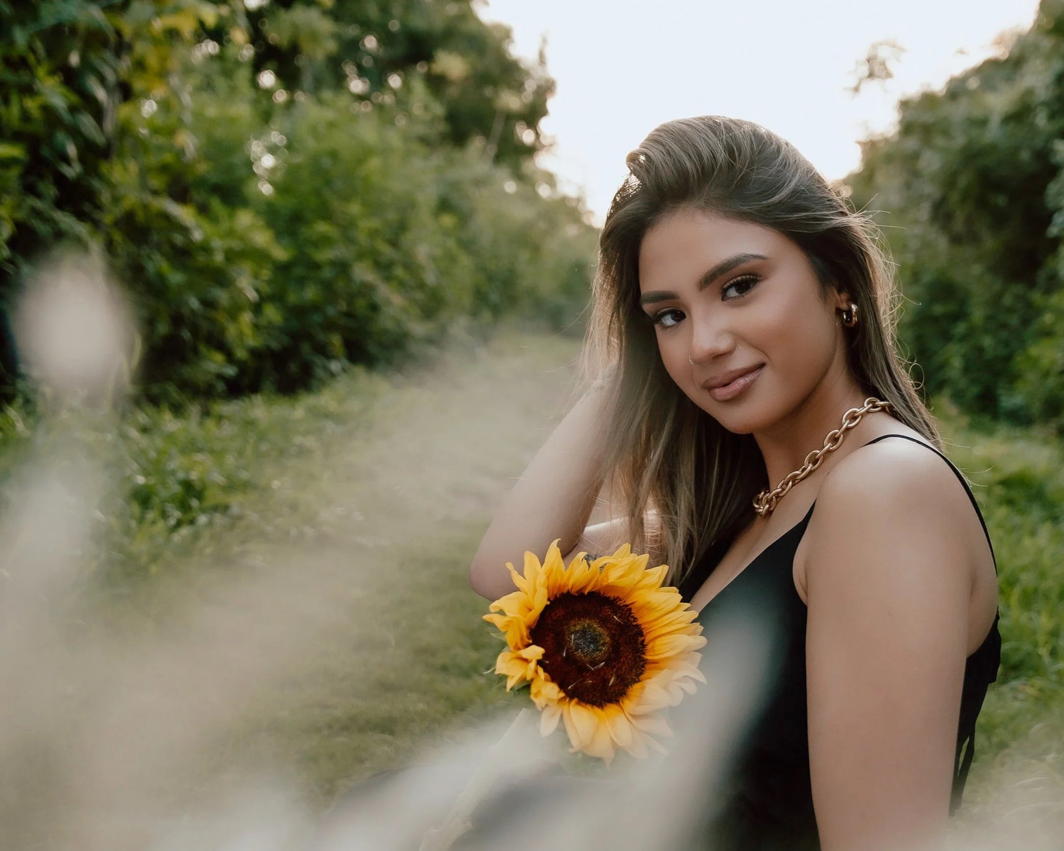 A young woman with long brown hair, wearing a black sleeveless top and gold jewelry, holding a sunflower while sitting outdoors on a grassy path with green trees in the background.