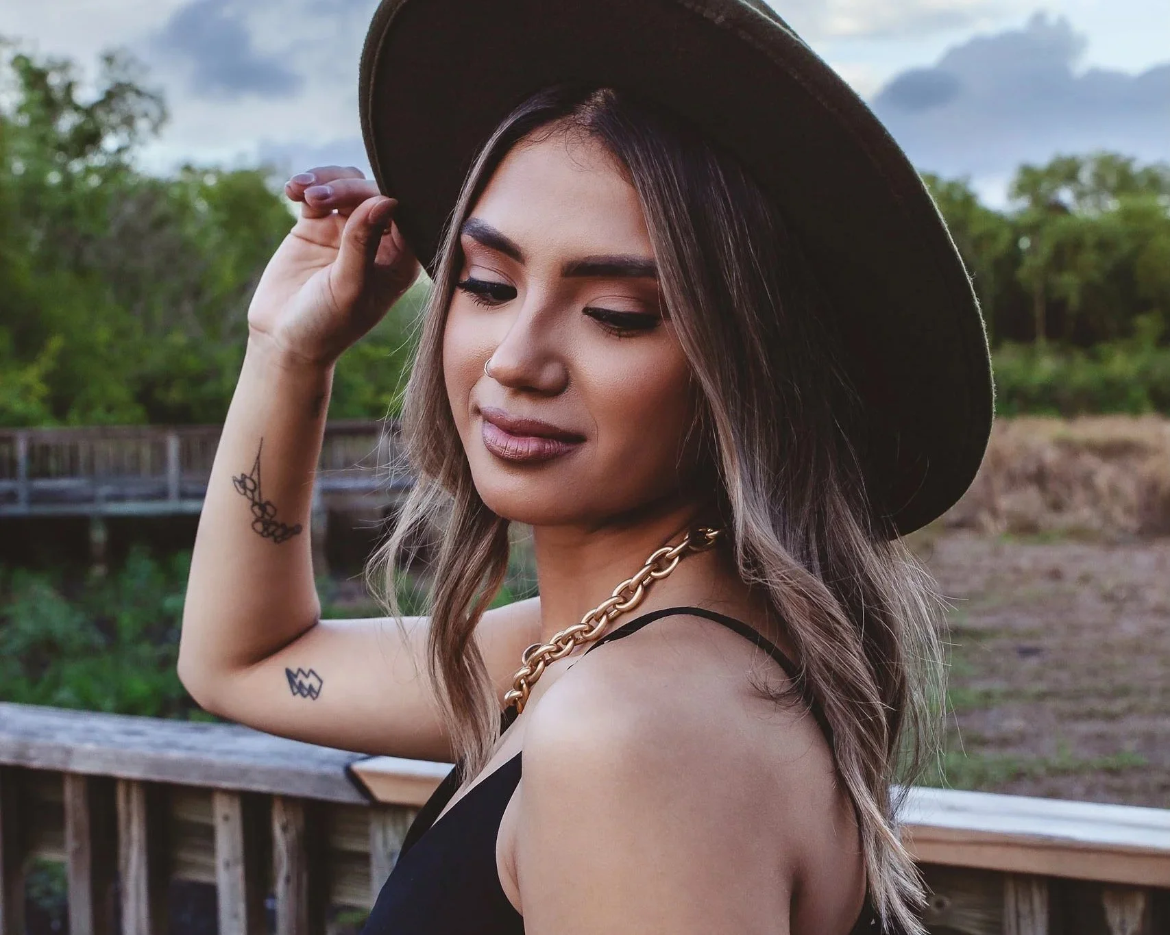 A young woman with light skin and tattoos on her arm, wearing a black dress, a chunky gold chain necklace, and a wide-brimmed hat. She is outdoors on a wooden deck with trees and a cloudy sky in the background, looking down and slightly smiling.