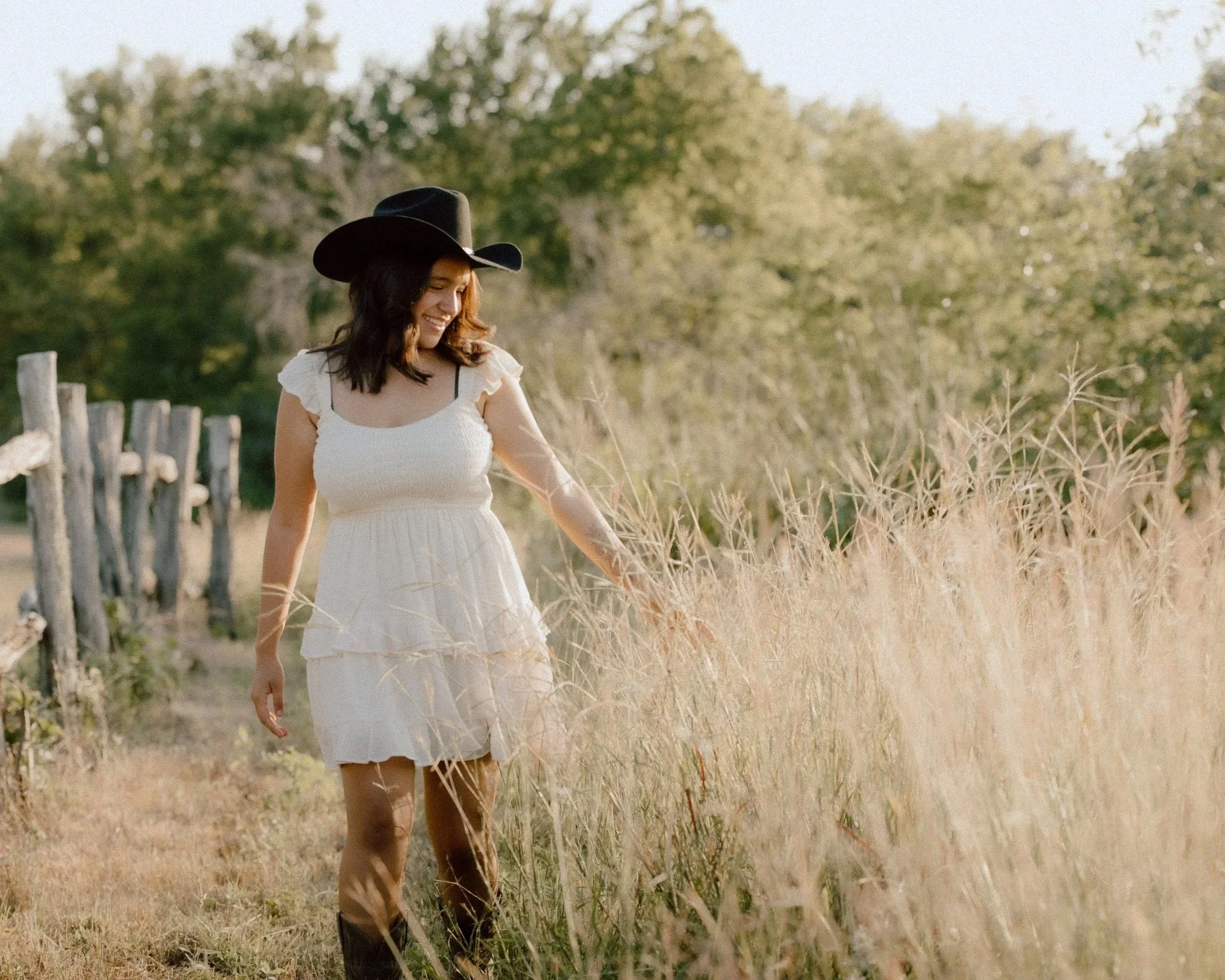 A woman in a white dress and a wide-brimmed black hat walking through a field of tall golden grass, smiling and looking down at the grass in a rural setting during daytime.