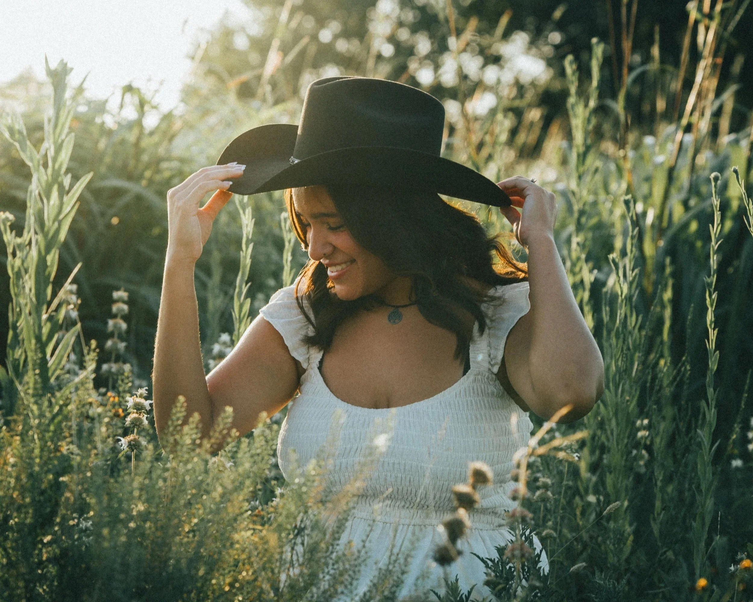 A woman wearing a white dress and a black wide-brimmed hat, holding the brim with both hands, smiling and looking down in a lush green field during sunset.