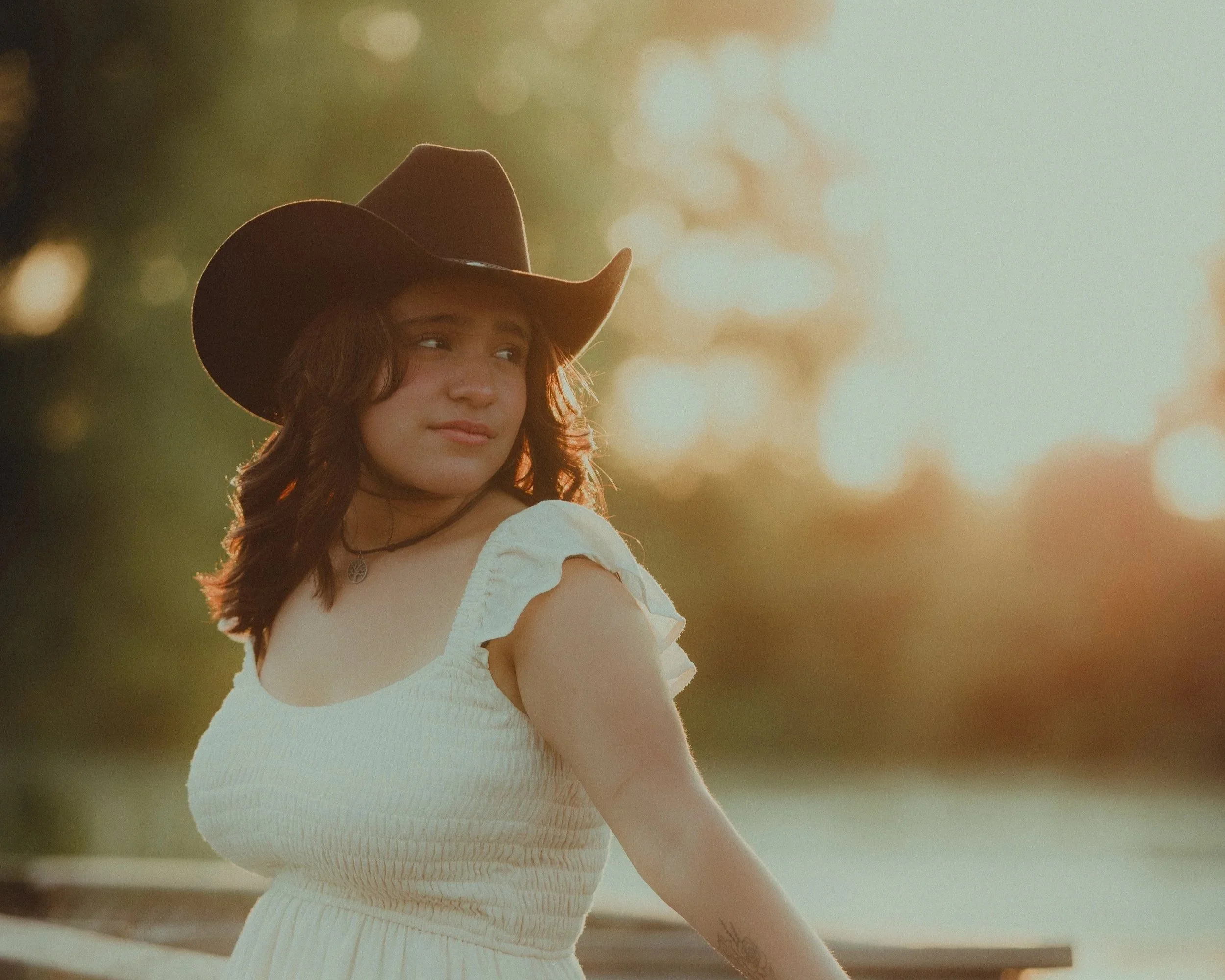 A young woman wearing a large cowboy hat and a white dress, looking to her left, standing outdoors during sunset with warm lighting.