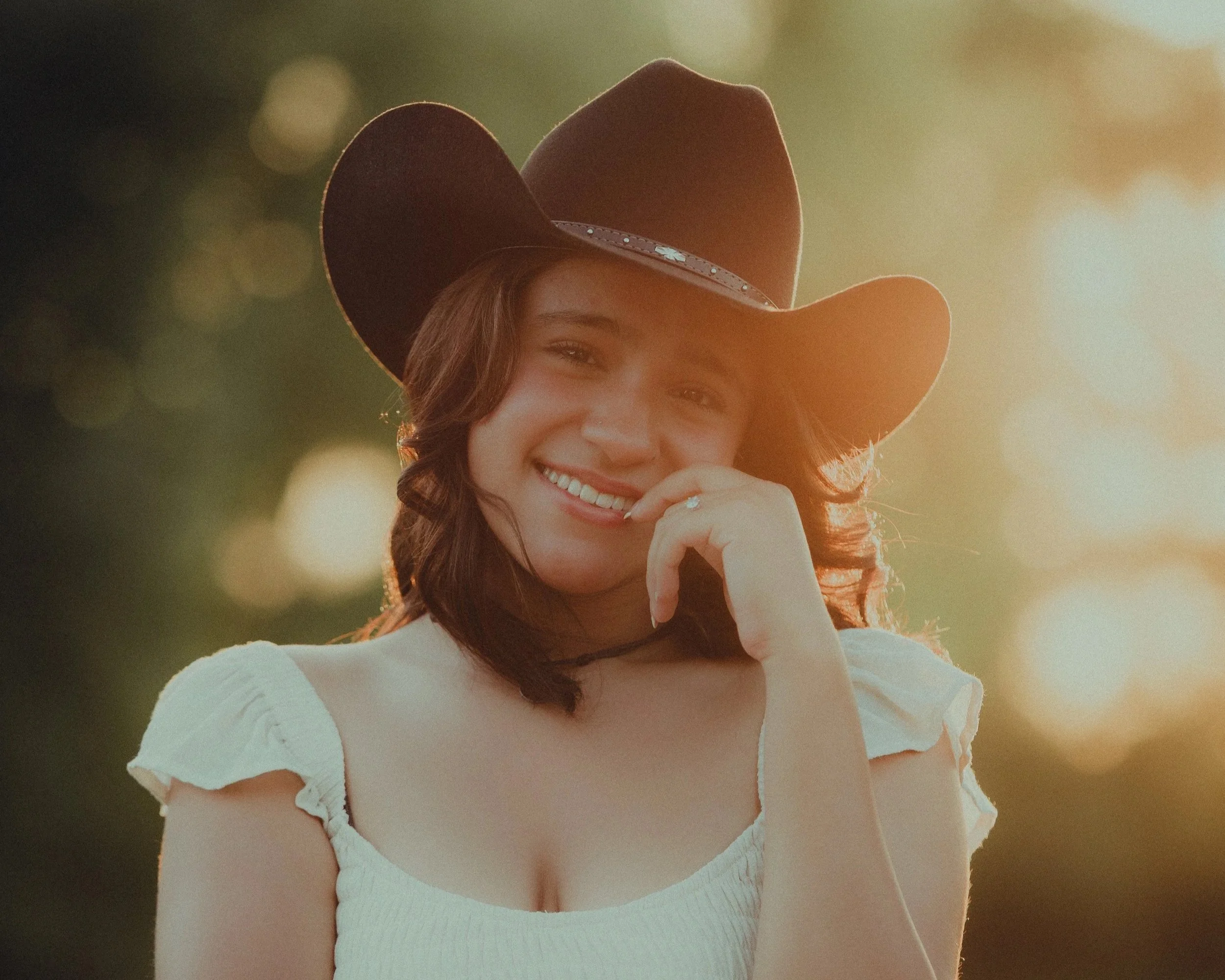 A young woman with brown hair smiles while wearing a cowboy hat and a white top, standing outdoors during sunset.