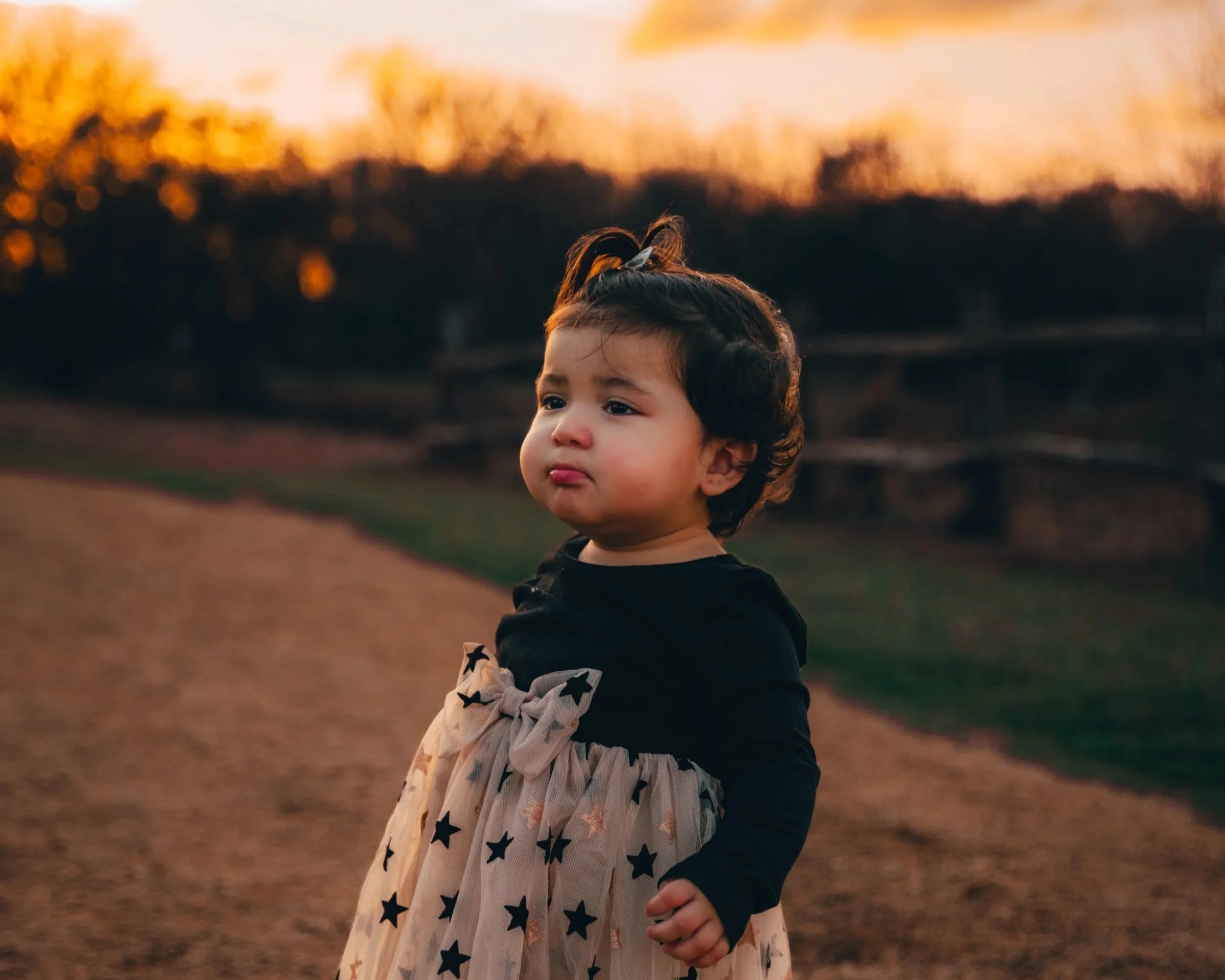 A young girl with dark hair and a top ponytail tied with a ribbon stands outdoors on a dirt path at sunset, wearing a black top and a beige skirt with black star patterns.