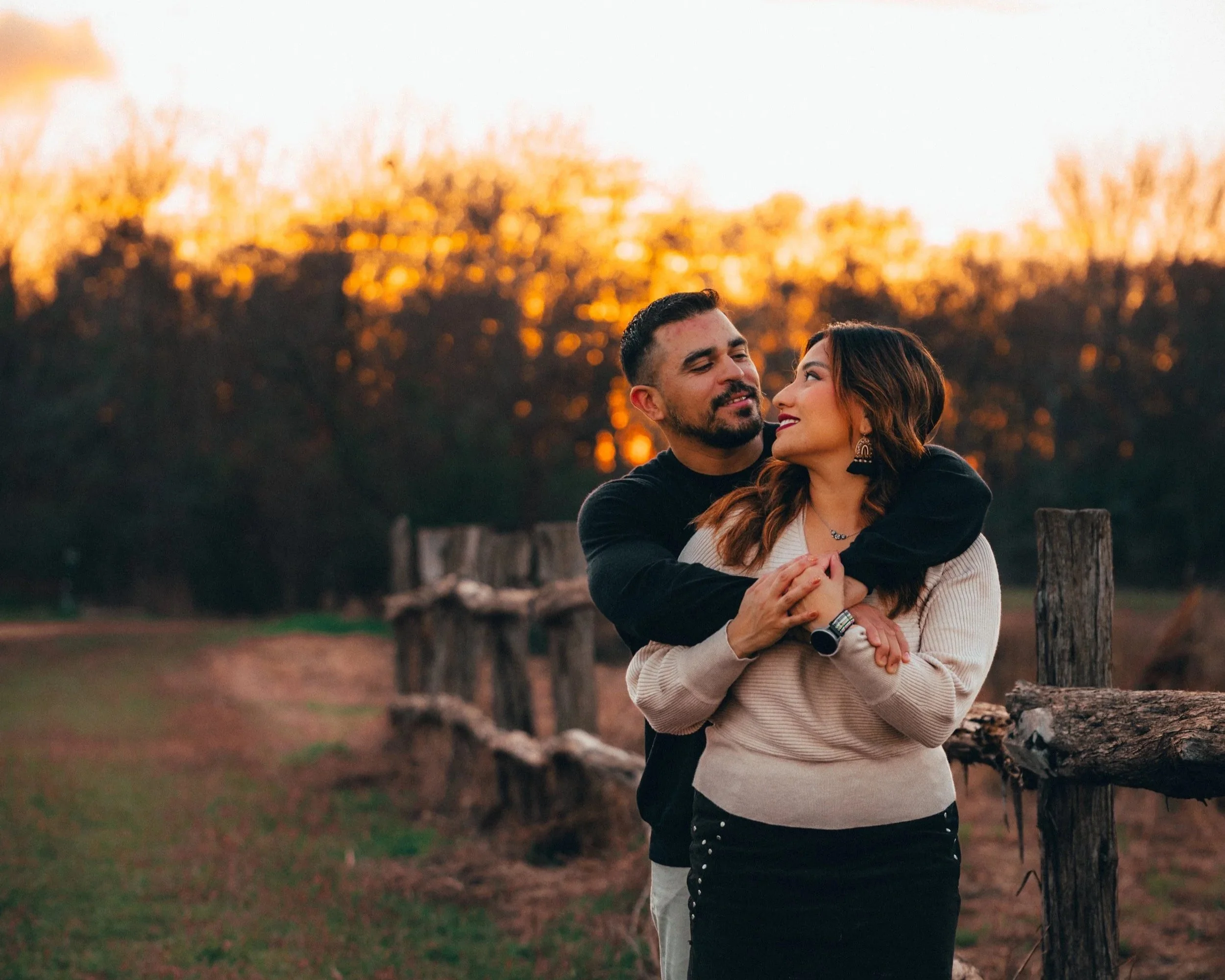 A couple embraces outdoors during sunset, with trees and a wooden fence in the background.