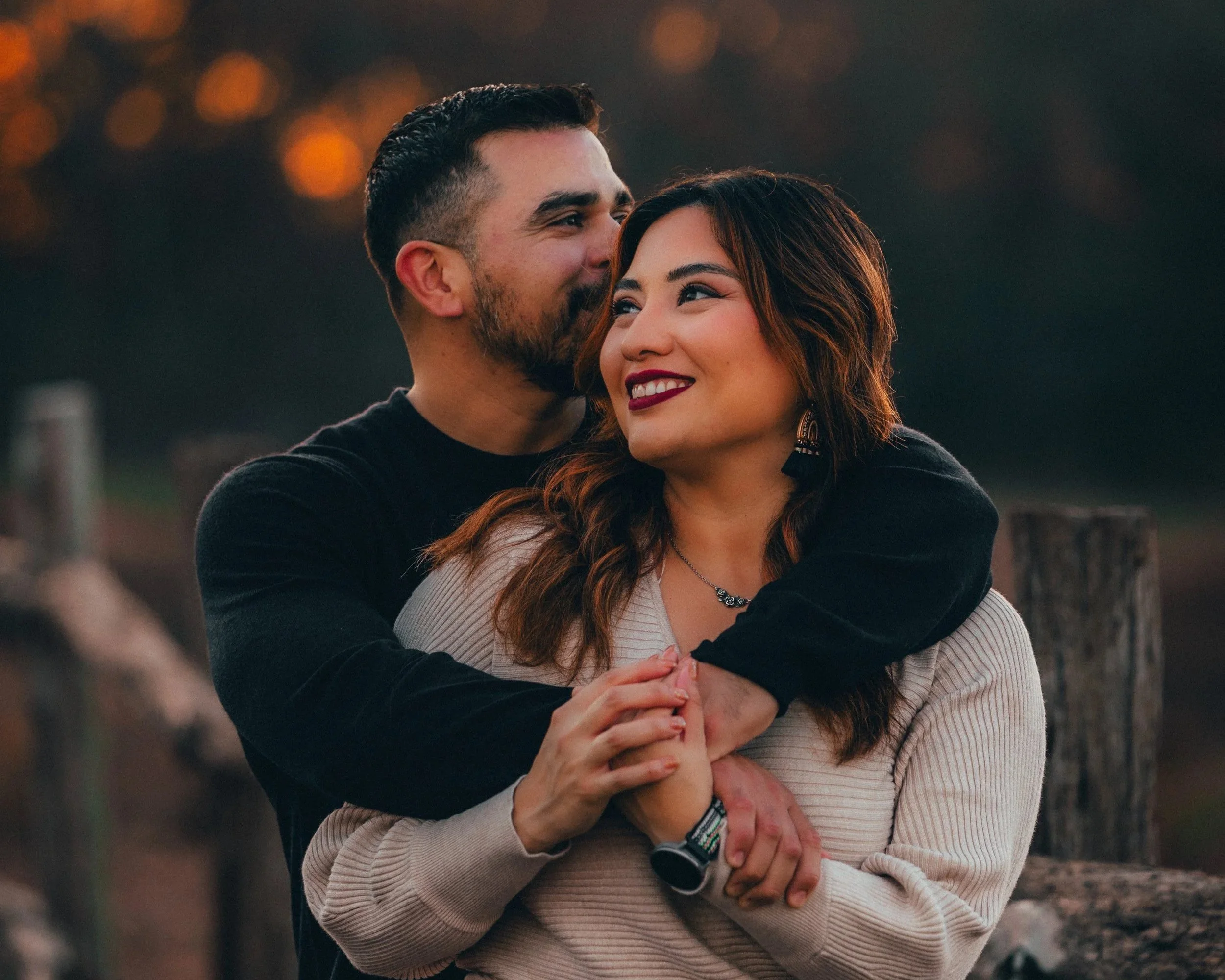 A happy couple standing outdoors during sunset, embracing each other and smiling with warm expressions.
