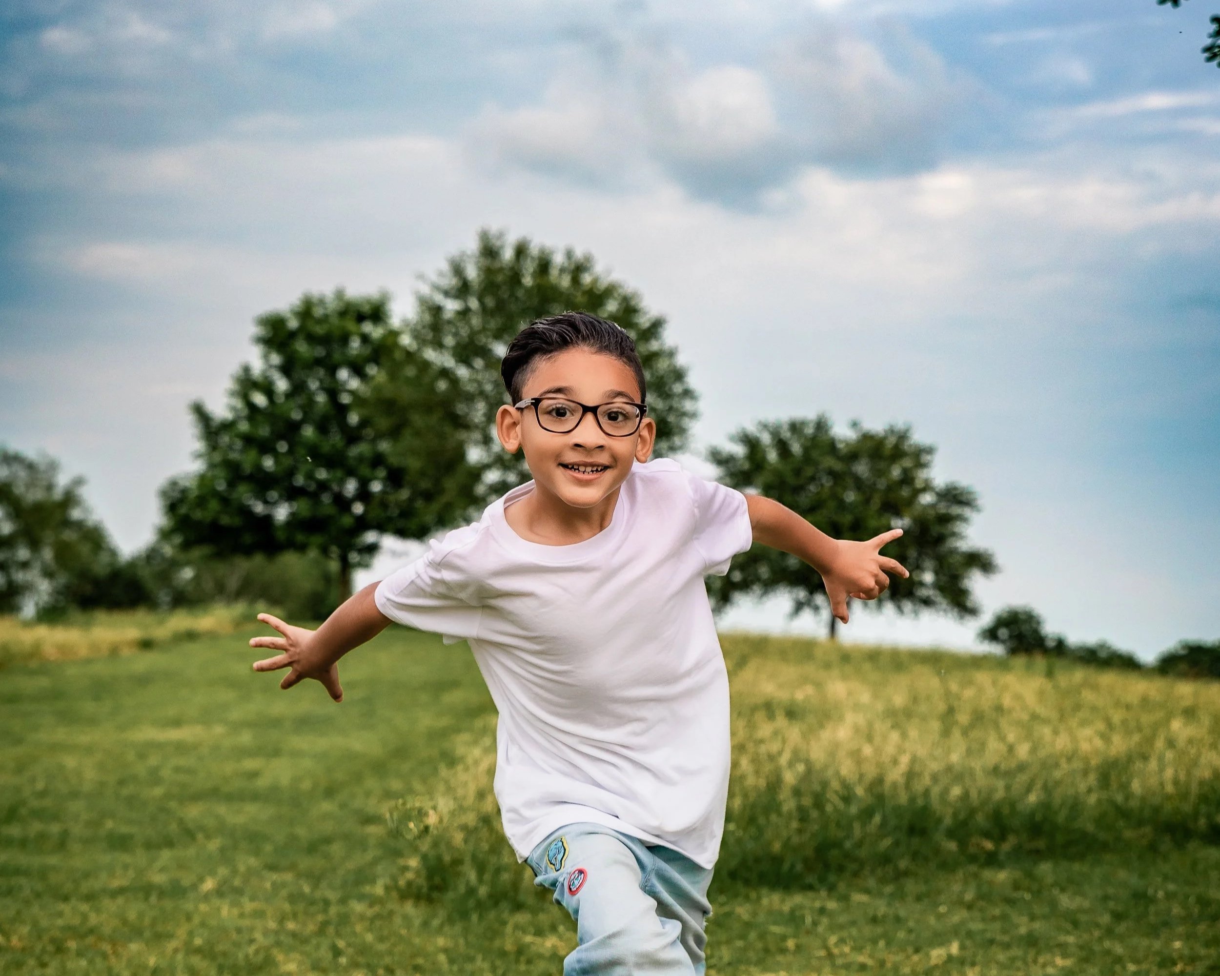 A young boy with glasses running happily in a grassy field with trees and a cloudy sky in the background.