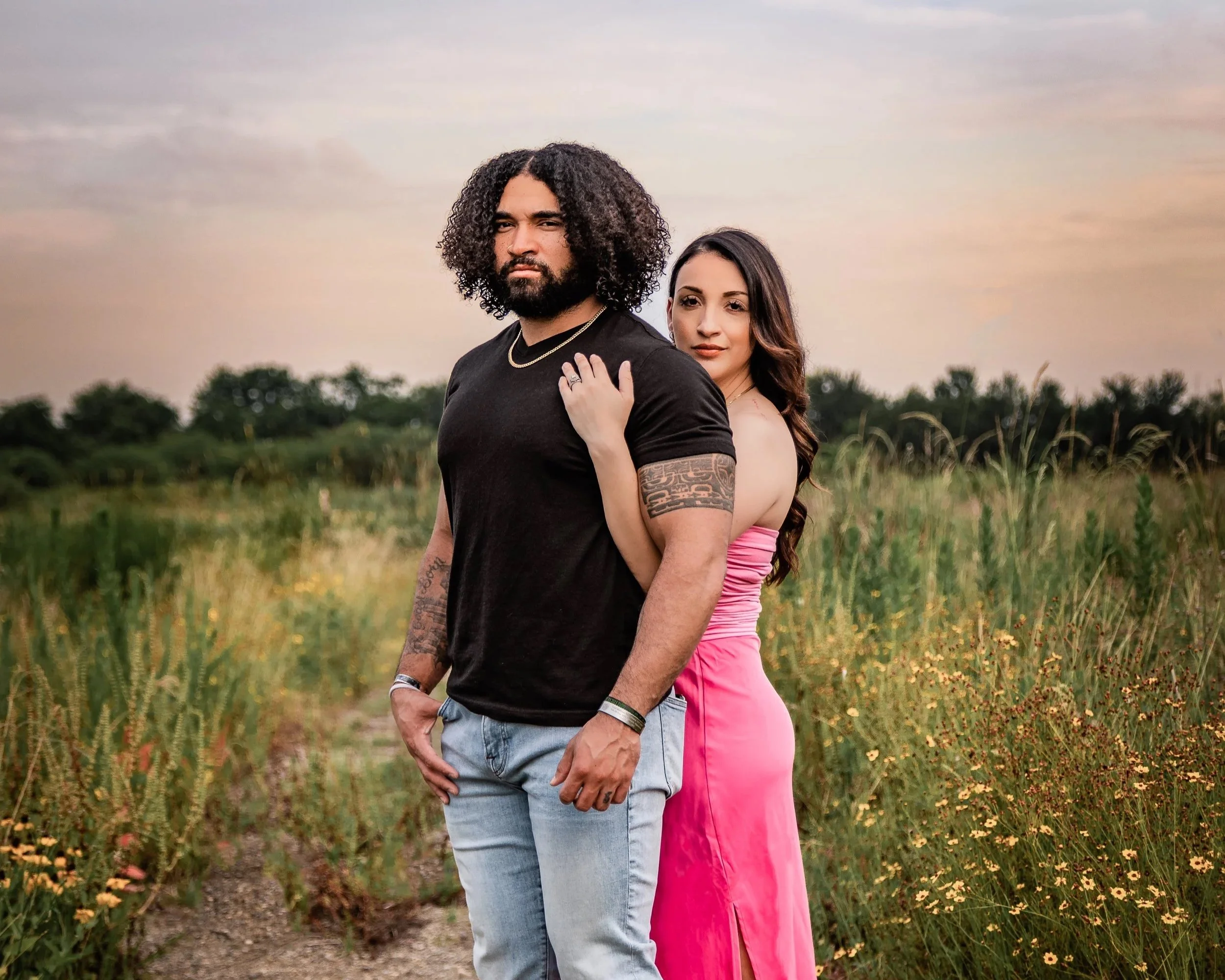 A man and woman standing together outdoors in a field at sunset, with the woman leaning on the man's shoulder.