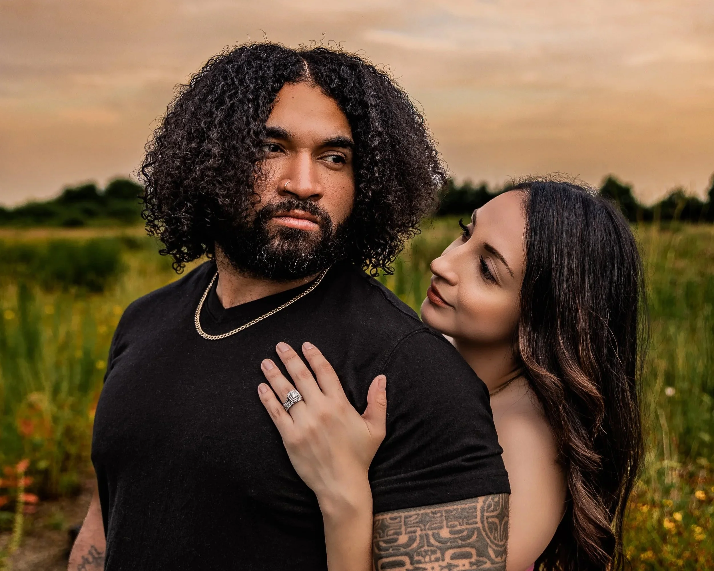 A man with curly hair and a beard stands outdoors at sunset, wearing a black shirt and silver jewelry. A woman with long dark hair and tattoos on her arm gently touches his shoulder, resting her head close to his. They are in a field with green plant