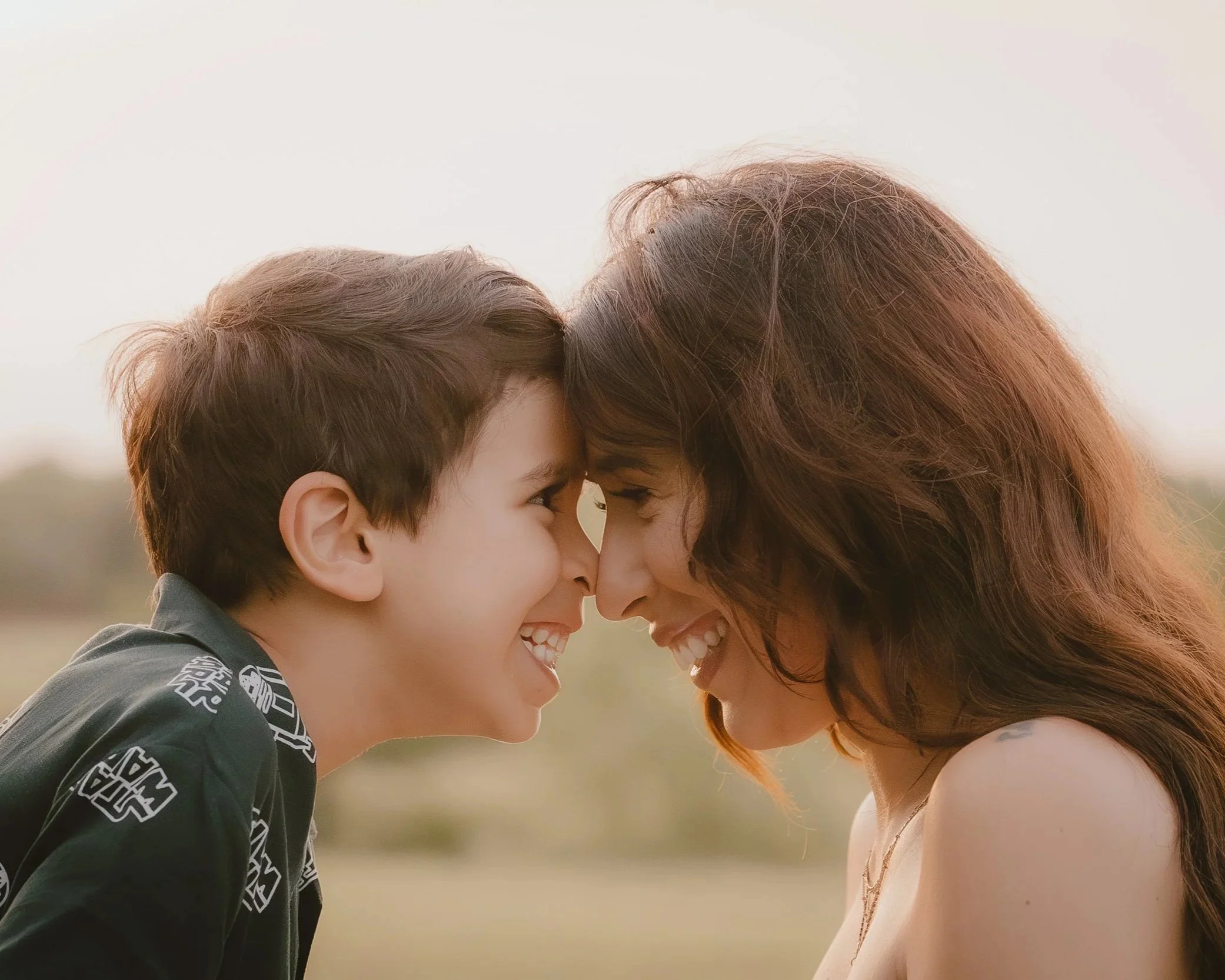 A young boy and a woman, likely his mother, smiling and touching foreheads outdoors during golden hour.