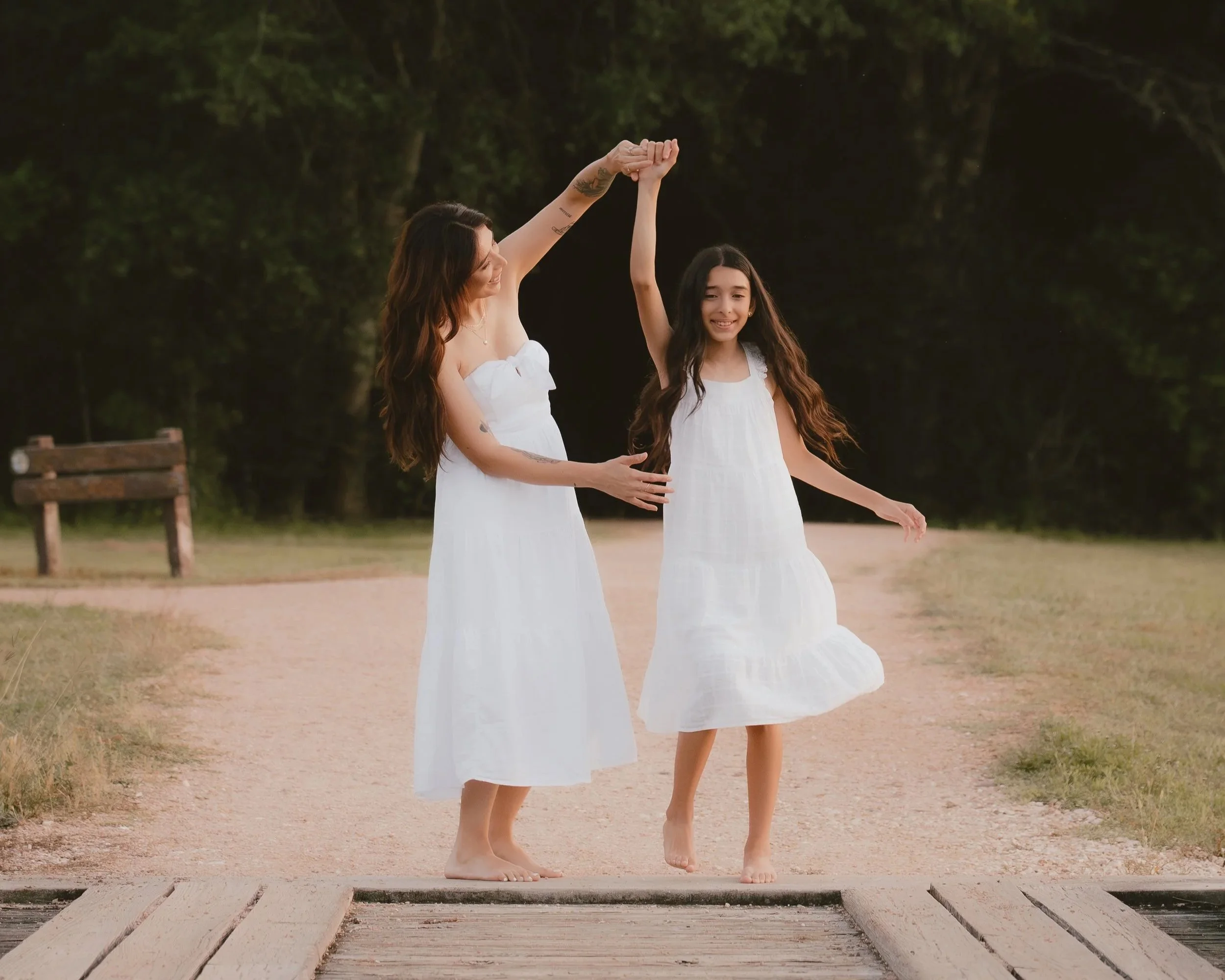 A woman and a girl in white dresses are dancing outdoors on a path at sunset. The woman is helping the girl spin, holding her hand up. Both are barefoot, with a park and trees in the background.