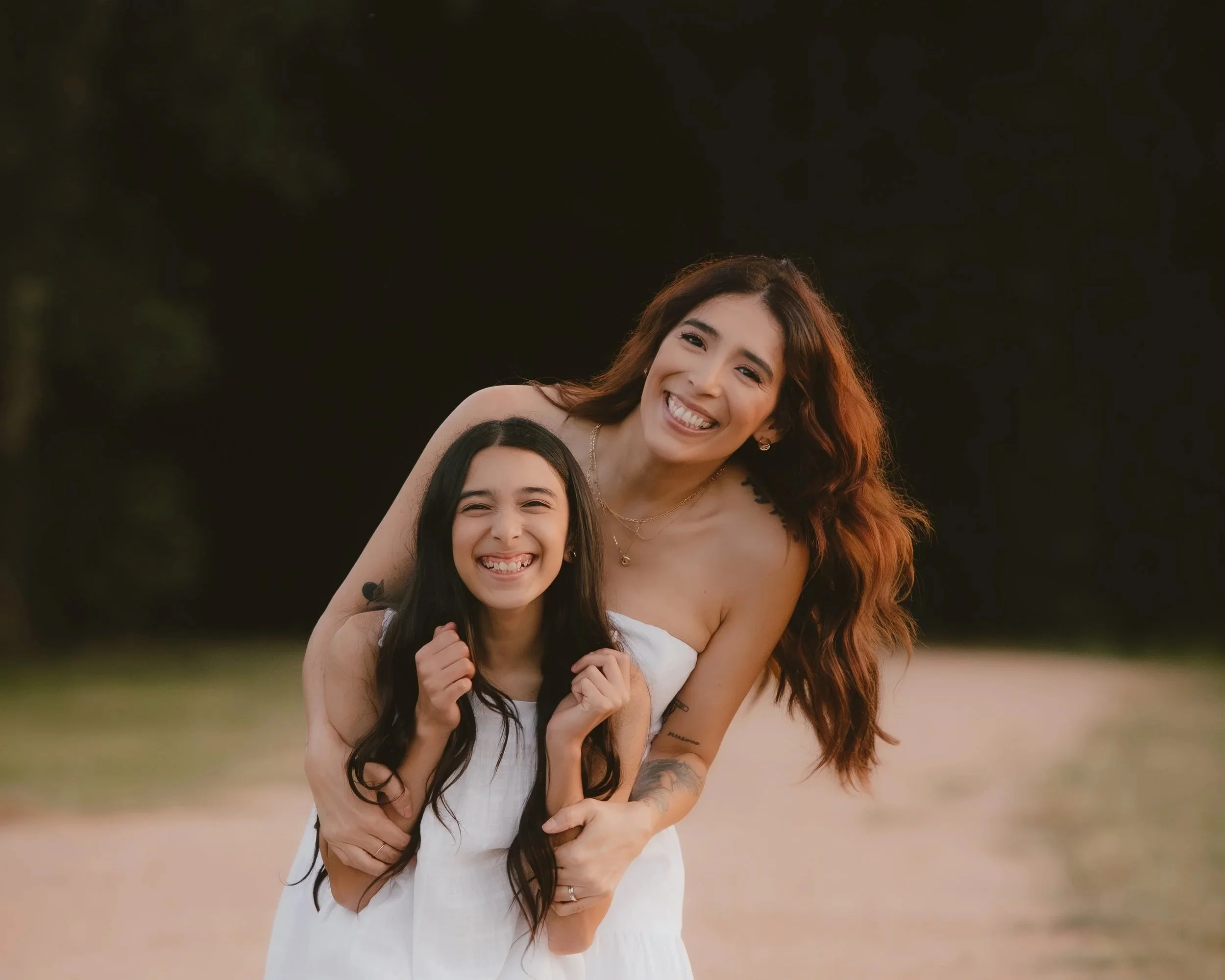A woman and a girl are smiling and hugging outdoors, both wearing white dresses, with a dark background and green grass.