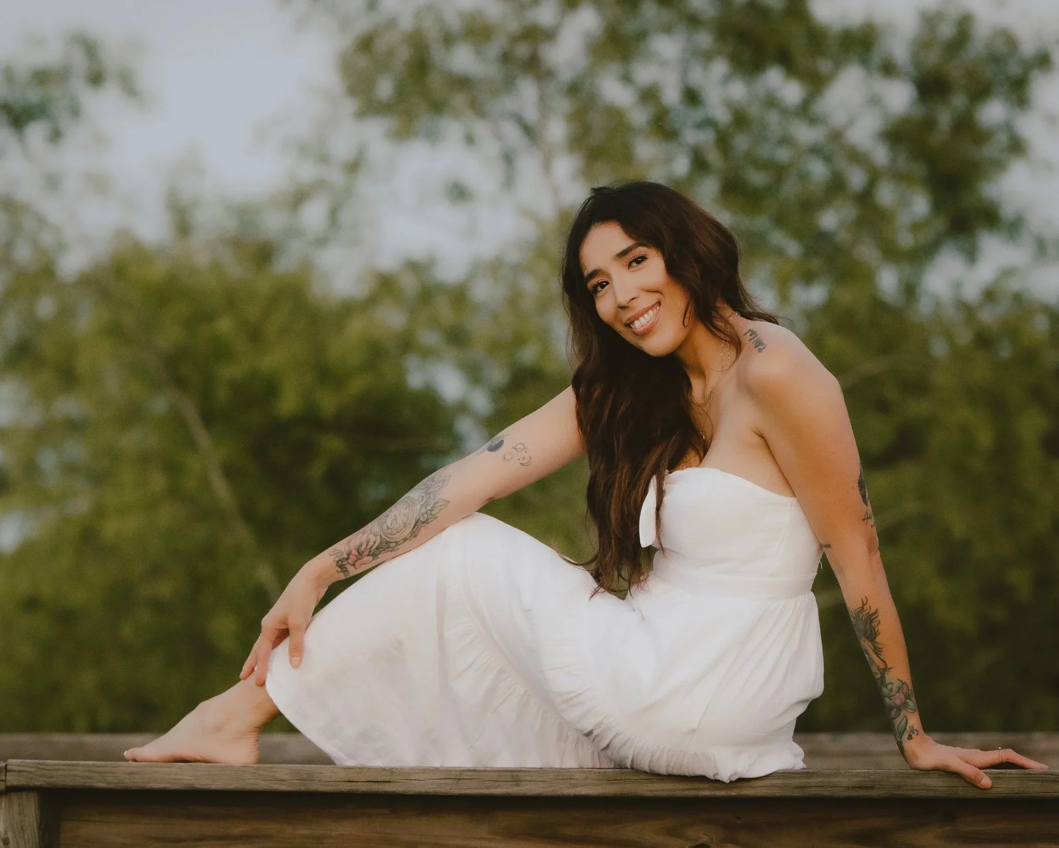 A young woman with long dark hair, tattoos on both arms, and wearing a white strapless dress, sitting on a wooden platform outdoors with trees in the background, smiling at the camera.
