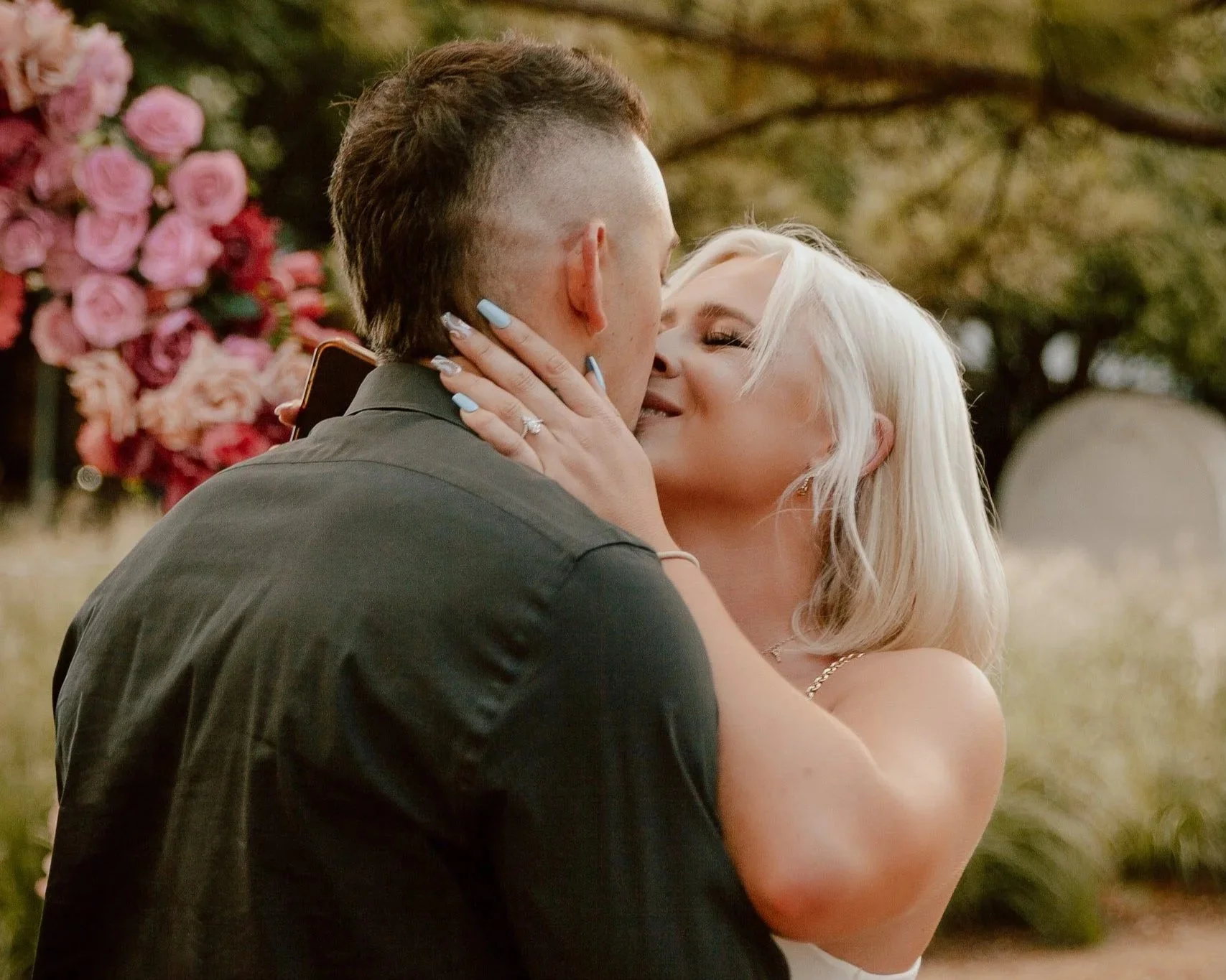 A couple sharing a kiss outdoors, with pink flowers and trees in the background.