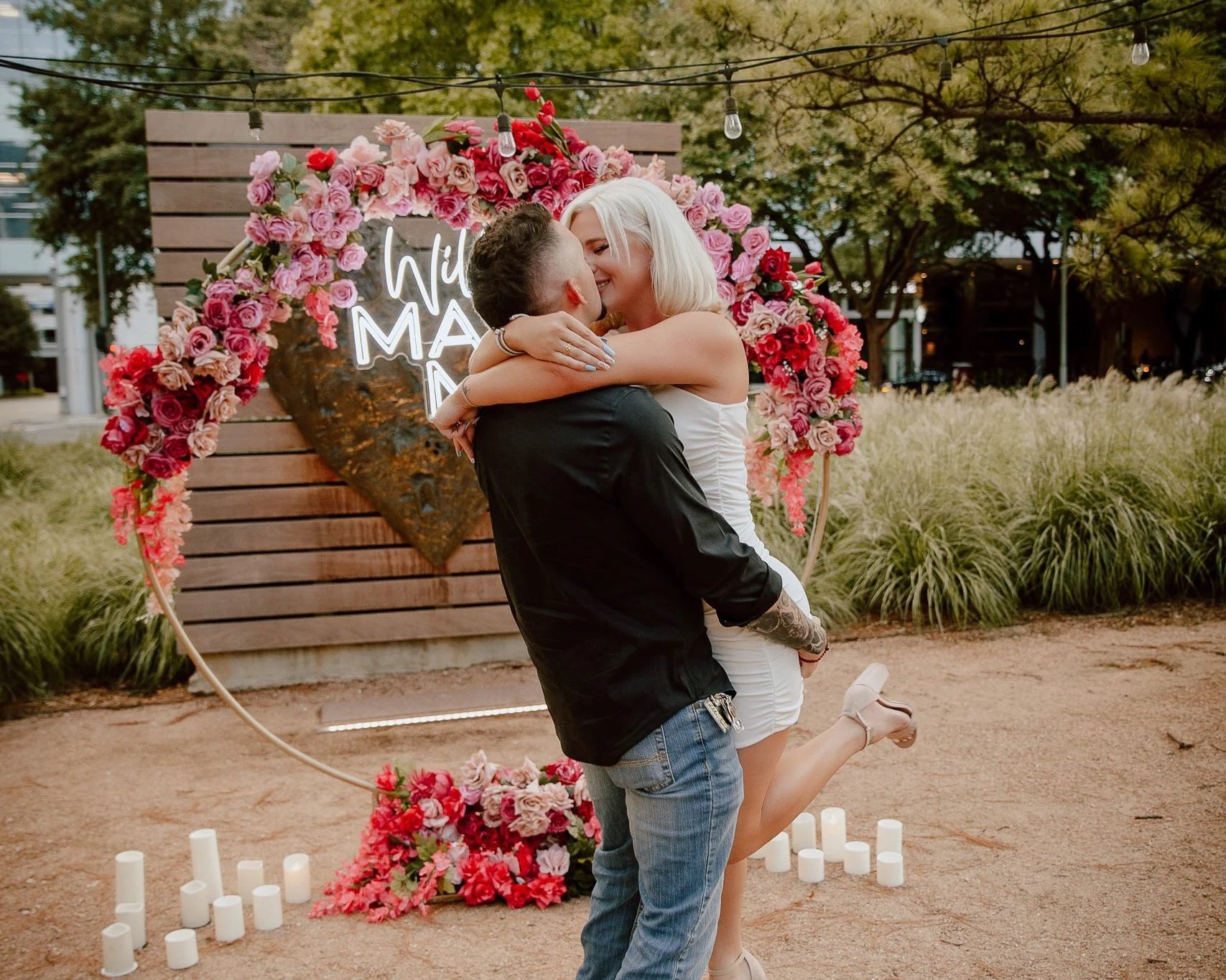 A couple embracing and kissing during a romantic outdoor event; woman has platinum blonde hair, and man has dark hair; decorated with a heart-shaped floral arch with pink and red roses, and a neon sign that reads a greeting; candles on ground