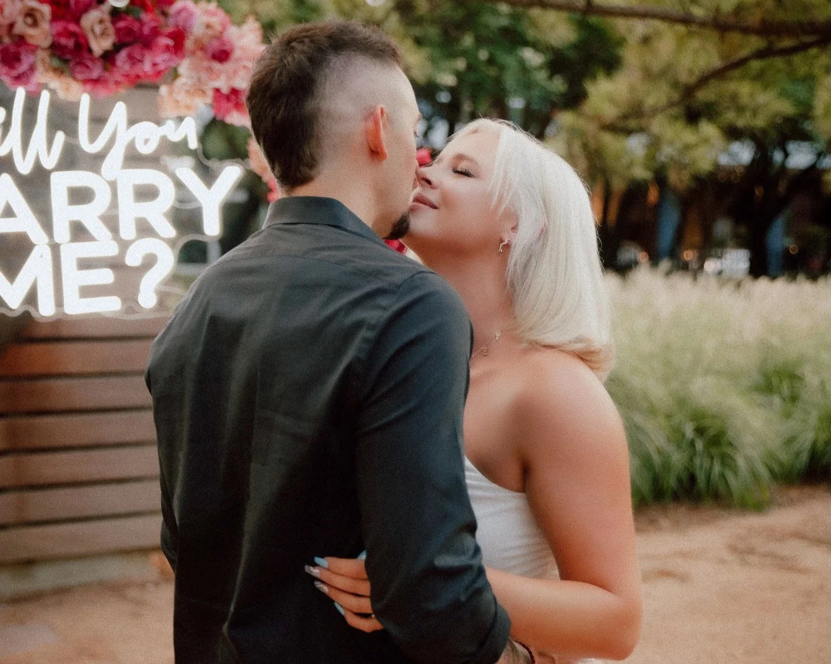 A couple is sharing a romantic moment outdoors at sunset, with the woman leaning in for a kiss, in front of a decorative sign asking 'Will You Marry Me?'. The woman has blonde hair and is wearing a white dress, while the man has dark hair styled in a