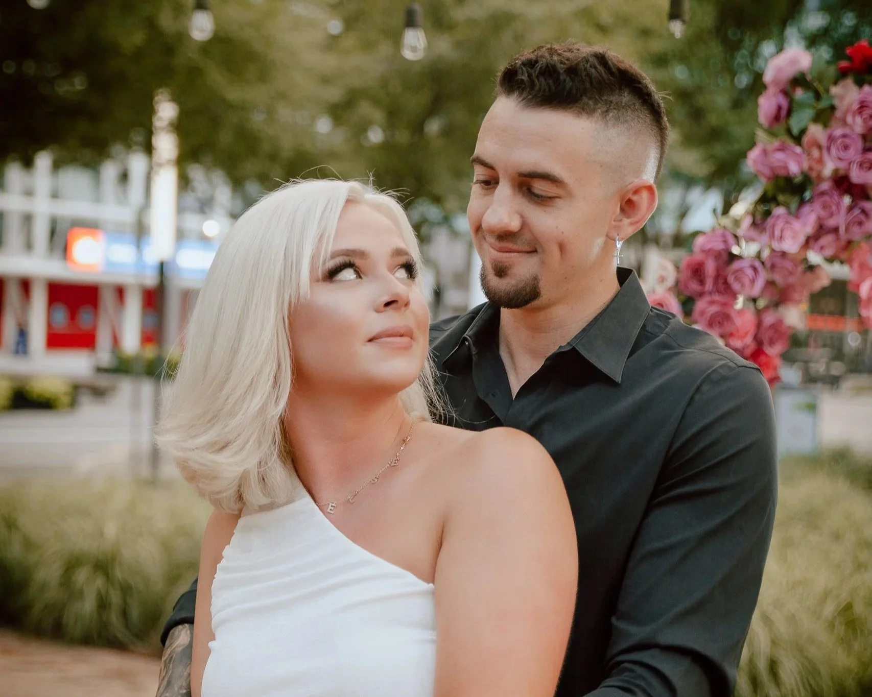 A young woman with short blonde hair and a white dress looking up at a young man with dark hair and a beard, who is smiling and looking at her. They are outdoors, with pink roses and string lights in the background.