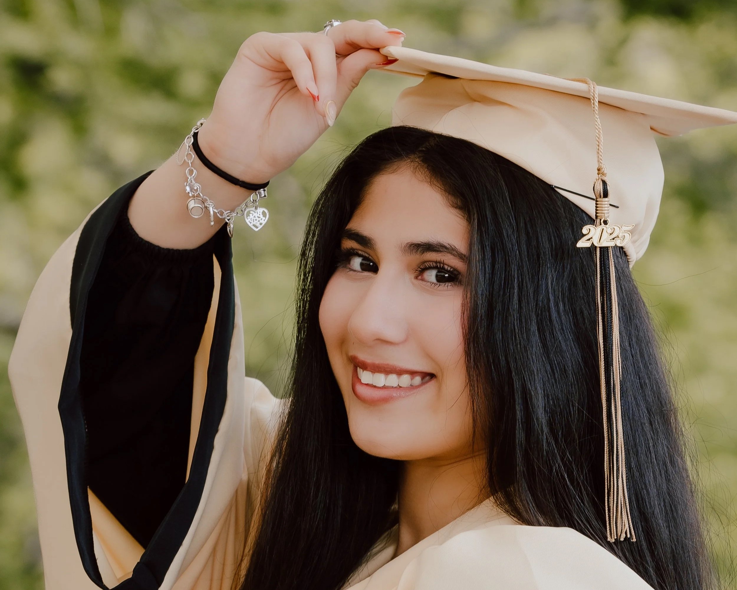 Young woman in a graduation cap and gown, smiling outdoors, holding the cap's tassel, with a green leafy background.