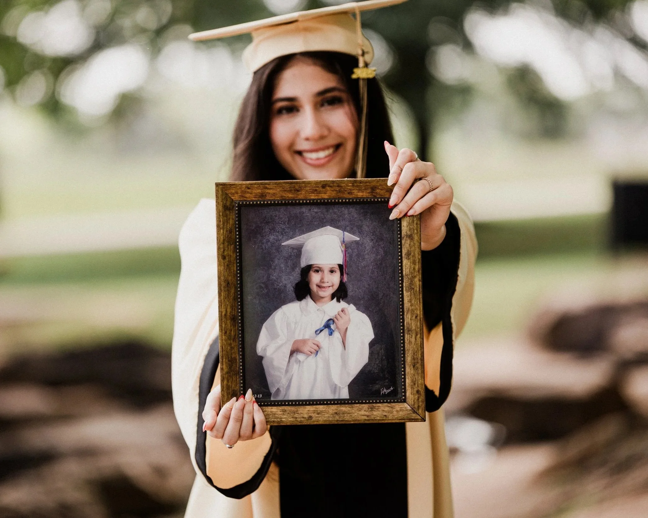 A young woman dressed in graduation cap and gown holding a framed photograph of herself as a child in graduation attire.