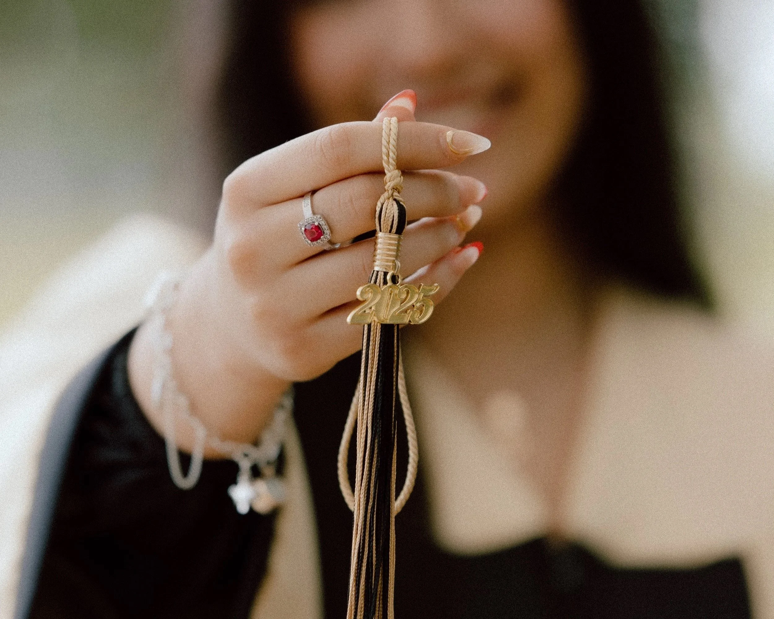 A woman holding a beige and black rosary with a gold 2025 charm. She is wearing a silver ring with a red gemstone and a silver bracelet.