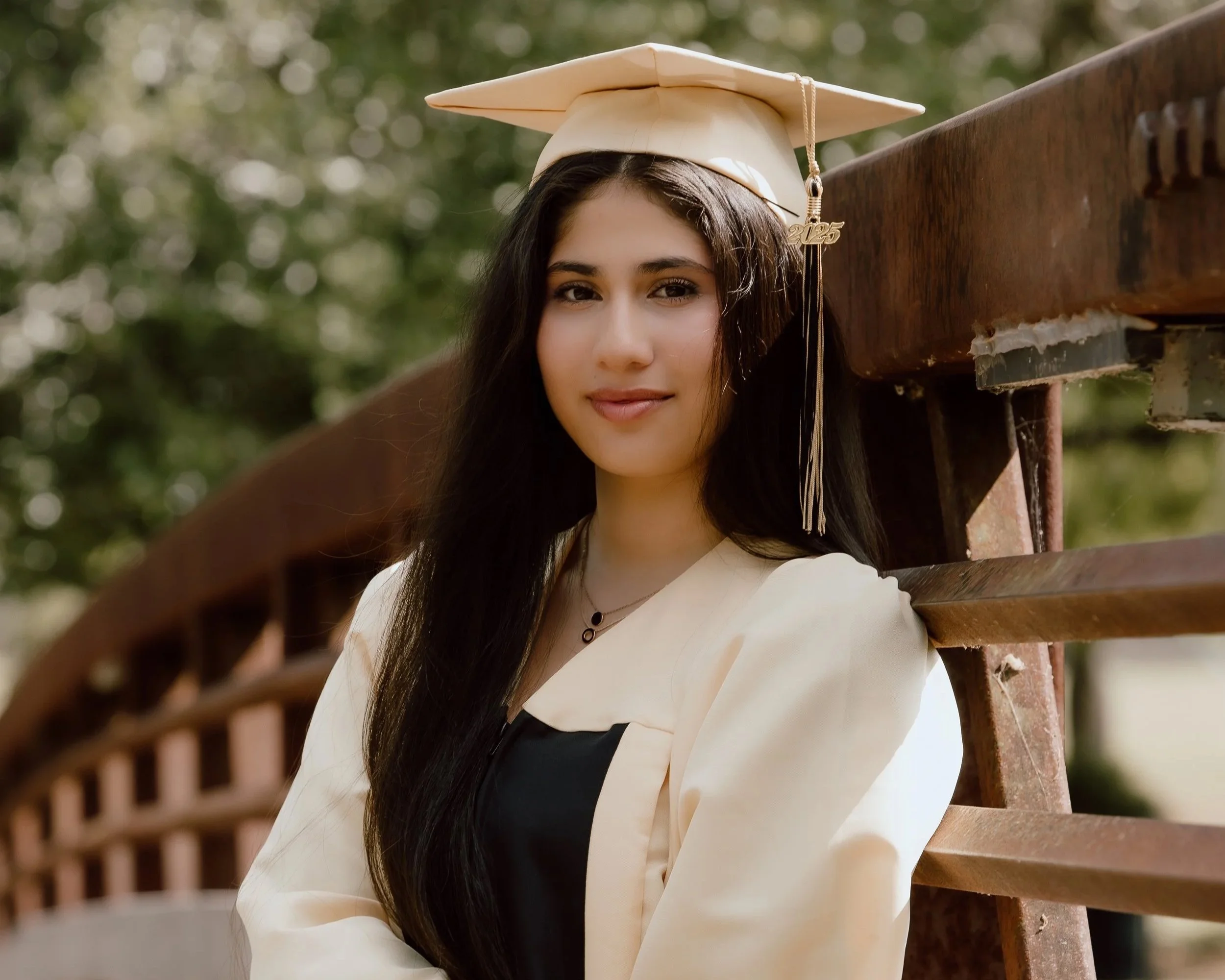 Young woman in a graduation cap and gown leaning against a wooden fence outdoors.