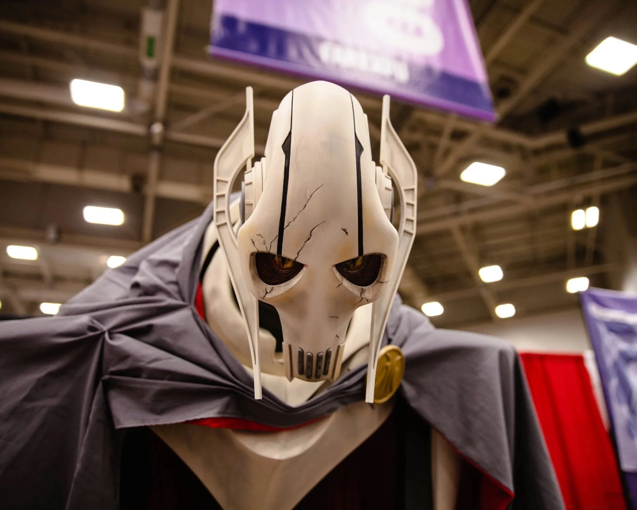 A person in a white skull mask with black cracks, yellow eyes, and a futuristic design, wearing a dark cape, standing indoors at a convention or event.
