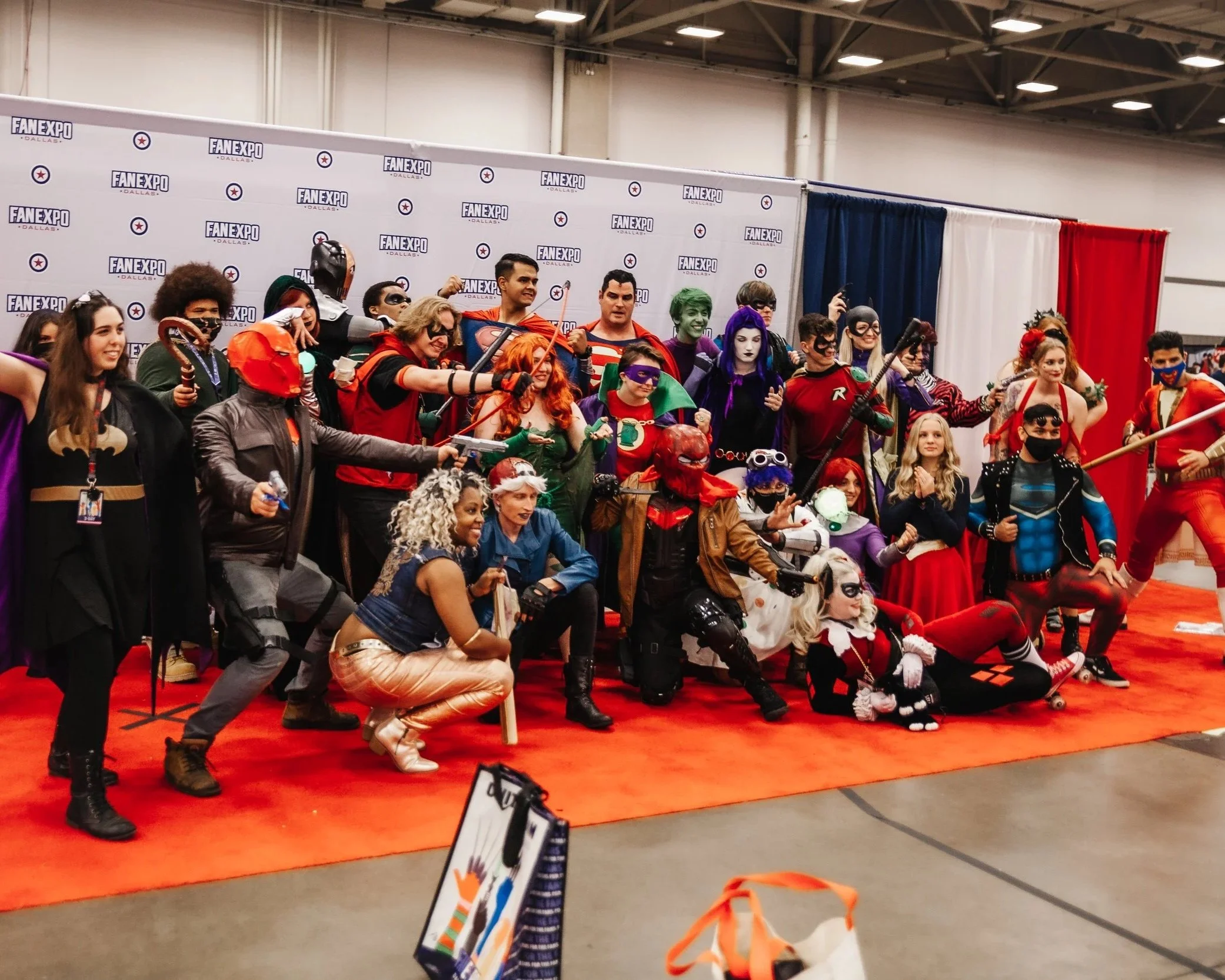 Group of people dressed as superheroes and comic book characters posing for a photo at a convention, with Batman, Robin, Harley Quinn, and others in costume, on a red carpet with a backdrop that says 'FAN EXPO Dallas'.