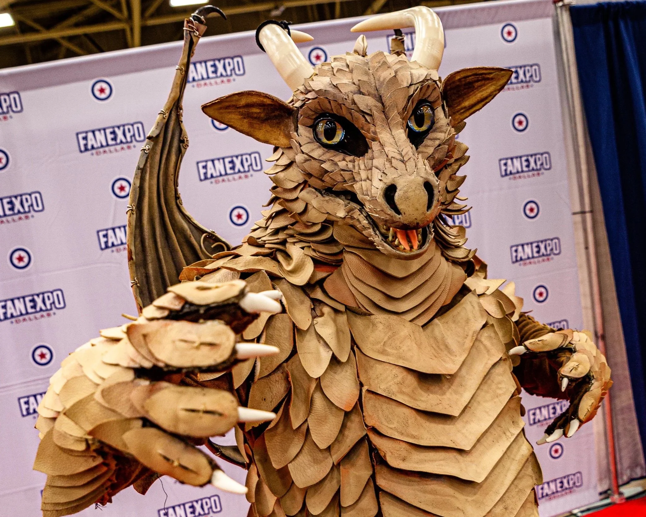 A person in a detailed dragon costume made of cardboard and other materials, with large eyes, horns, wings, and claws, at FanExpo Dallas.