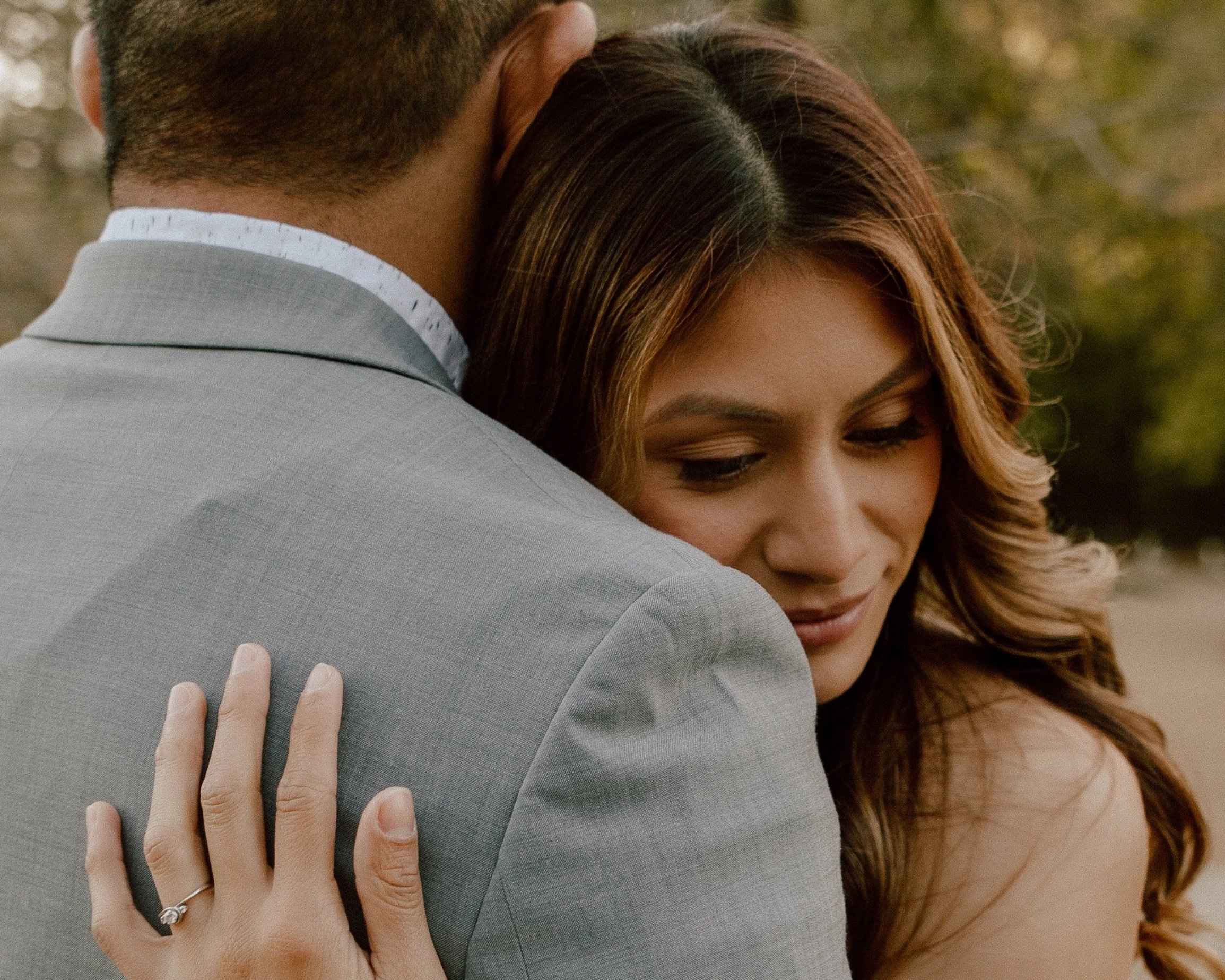 Close-up of a woman with long wavy hair embracing a man, her hand with an engagement ring resting on his shoulder. Both appear to be outdoors.