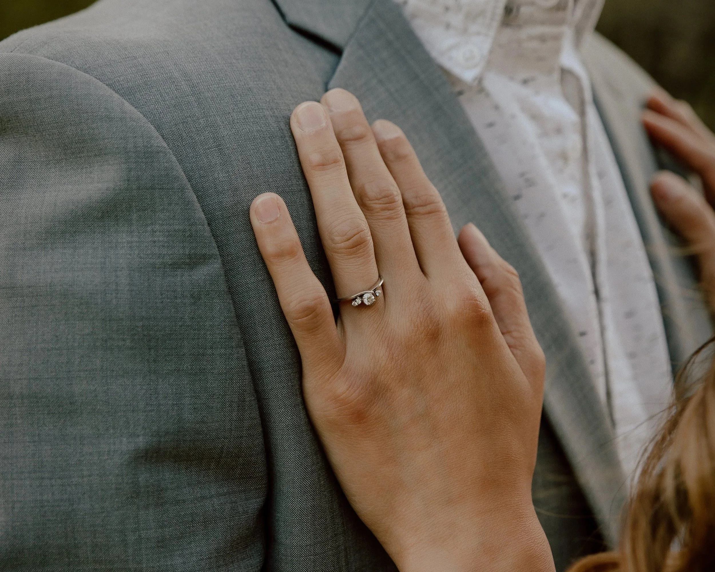 A woman wearing a ring with a large gemstone is resting her hand on a man's shoulder, who is dressed in a gray suit and a light-colored shirt.