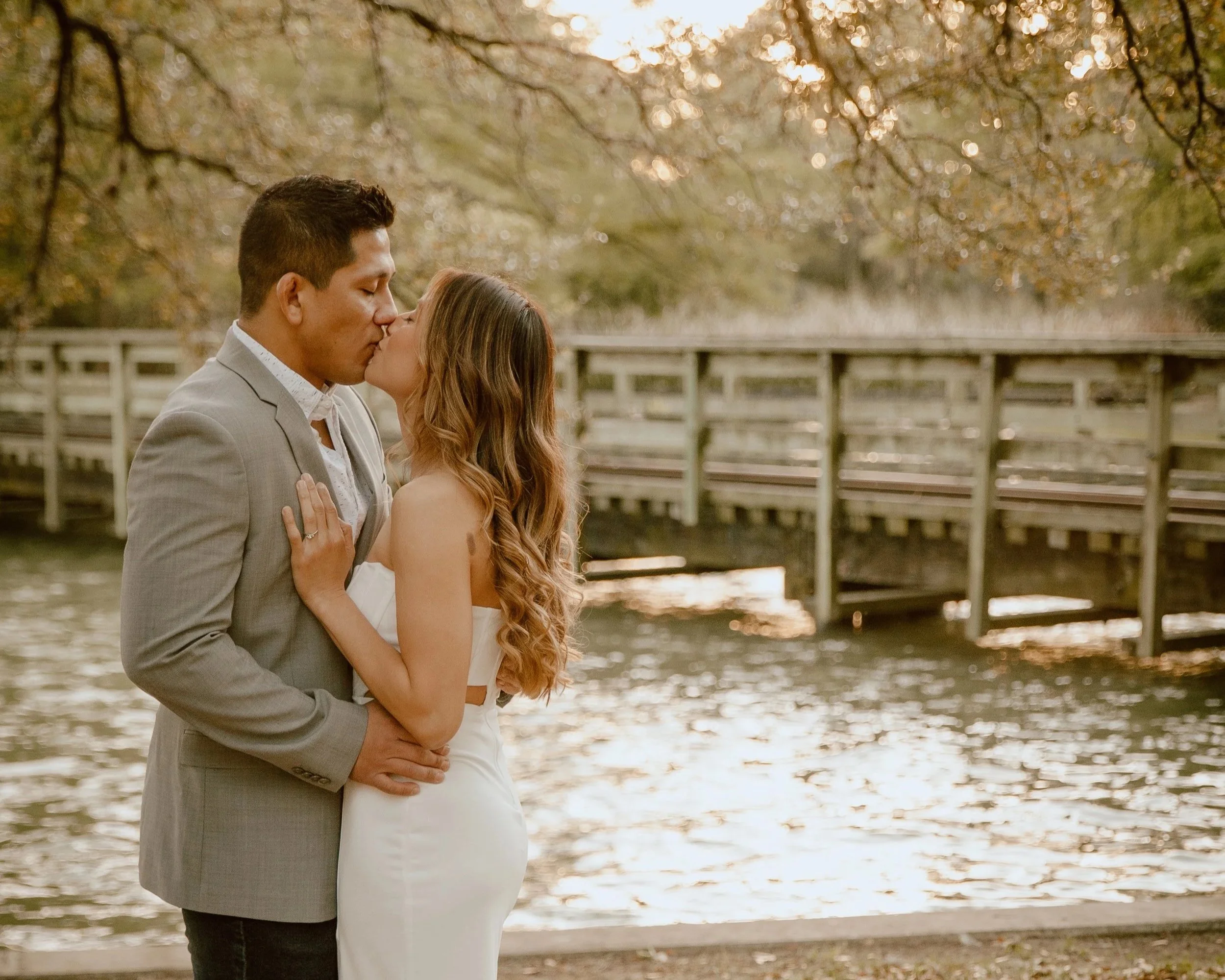A couple in wedding attire sharing a kiss by a river with a wooden bridge and autumn trees in the background.